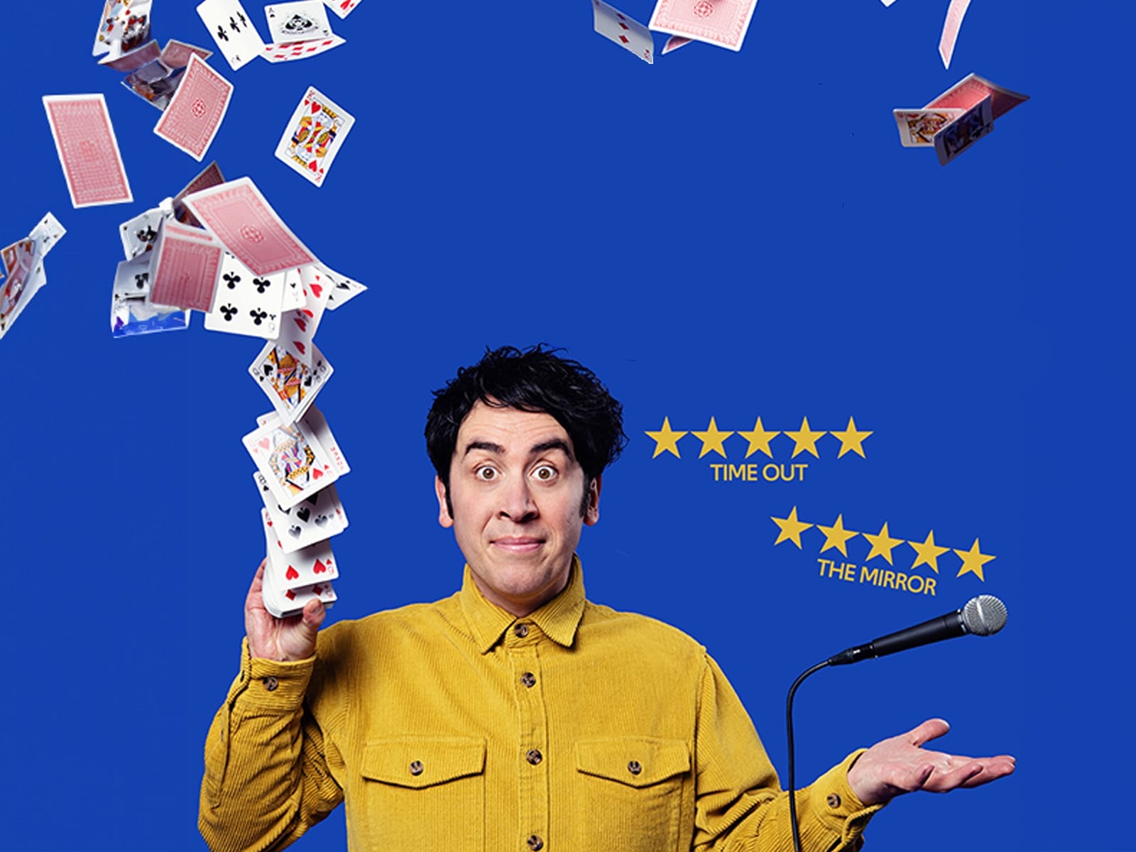 Comedian Pete Firman wears a yellow shirt, standing in front of a blue background having thrown cards in the air.