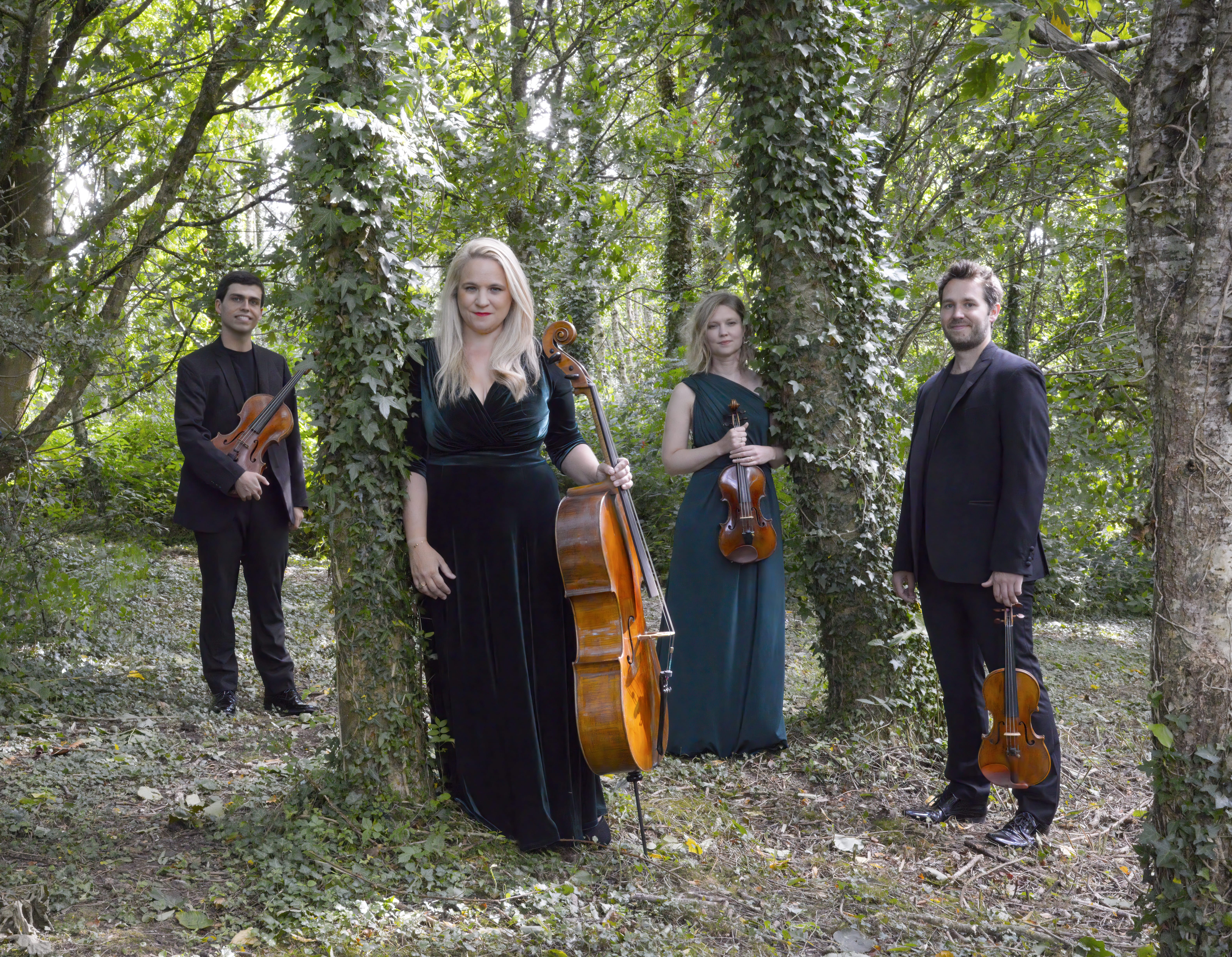a photo of four musicians holding their string instruments standing in a forest