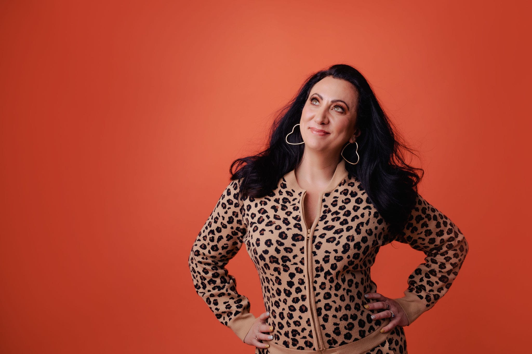 Comedian Alyssia Kyria stands in front of an orange background wearing a leopard print jumpsuit.