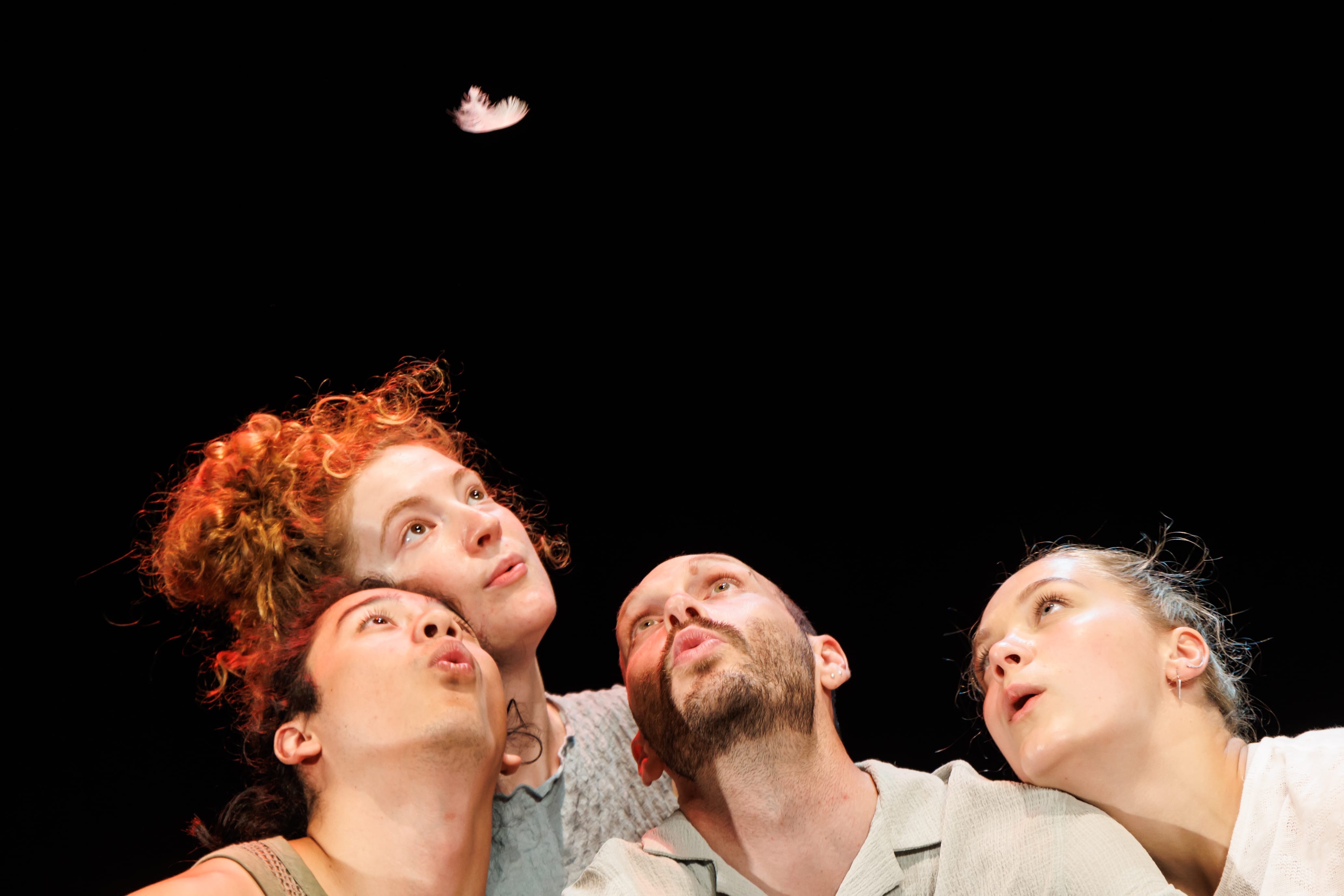 four people's heads are visible on the bottom of a photograph as they all look up and blow to keep a piece of feather in air together