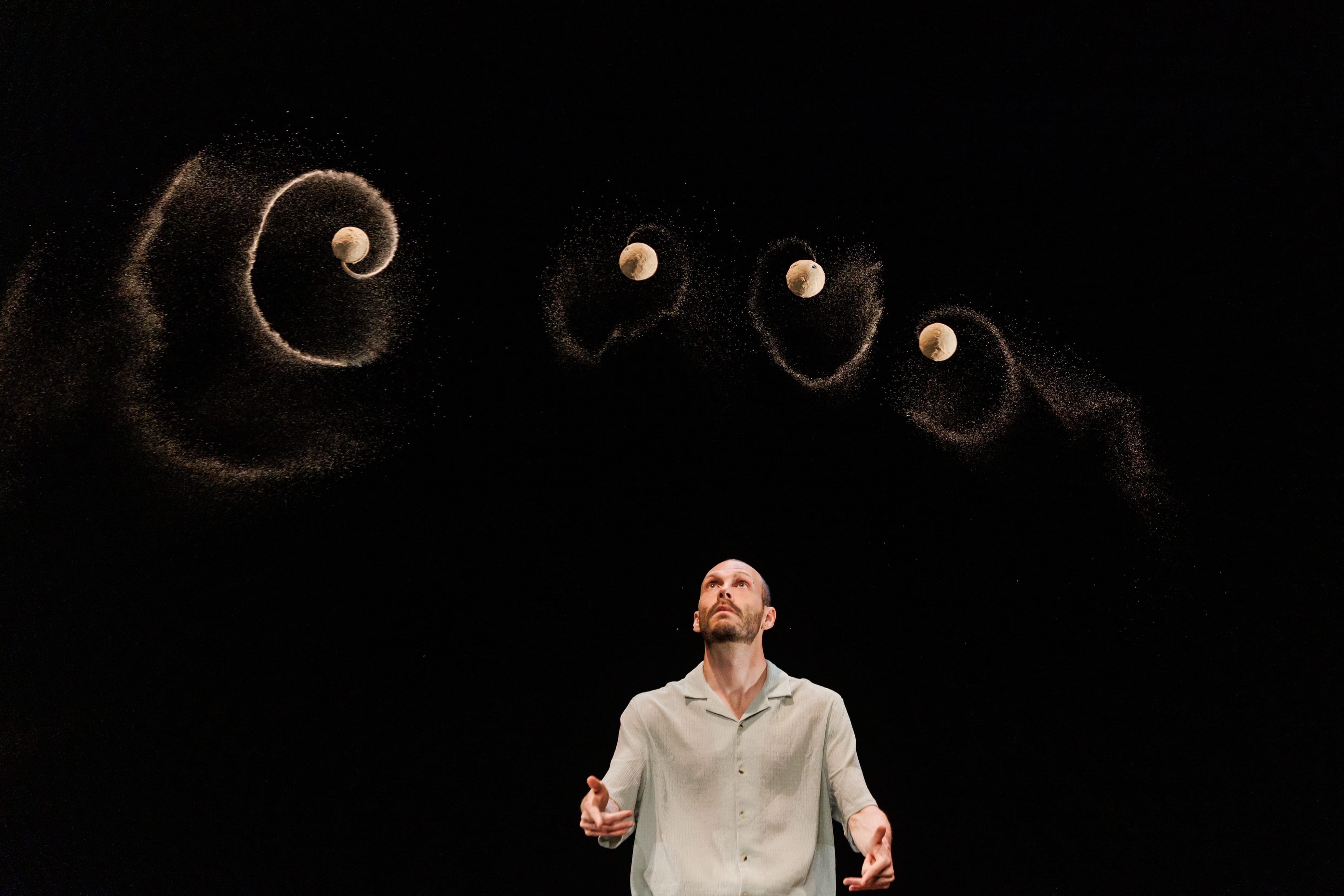 a man on a black stage wearing a light coloured shirt looks up while juggling a few balls