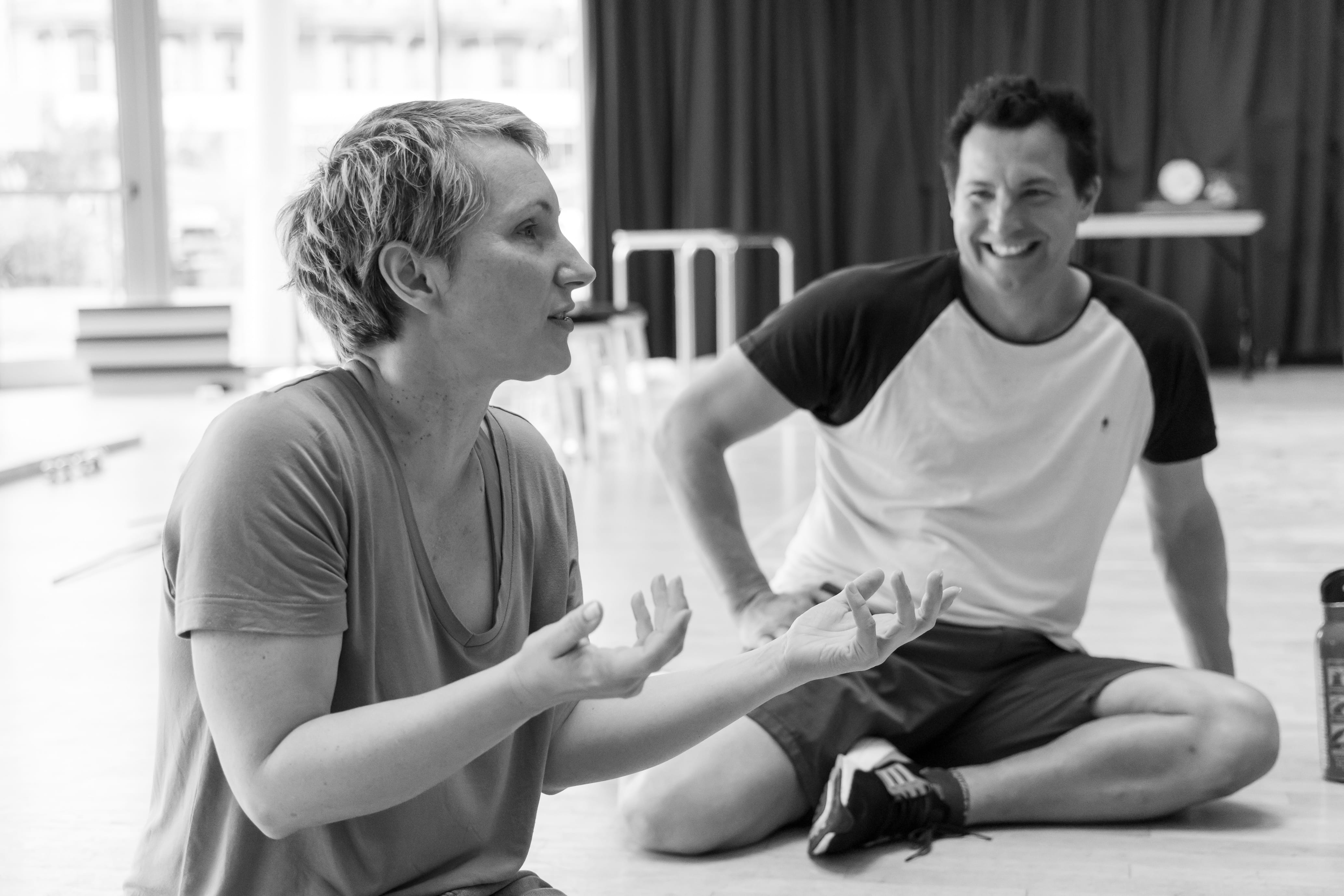 two people wearing comfortable clothes sit on the floor in a studio space chatting