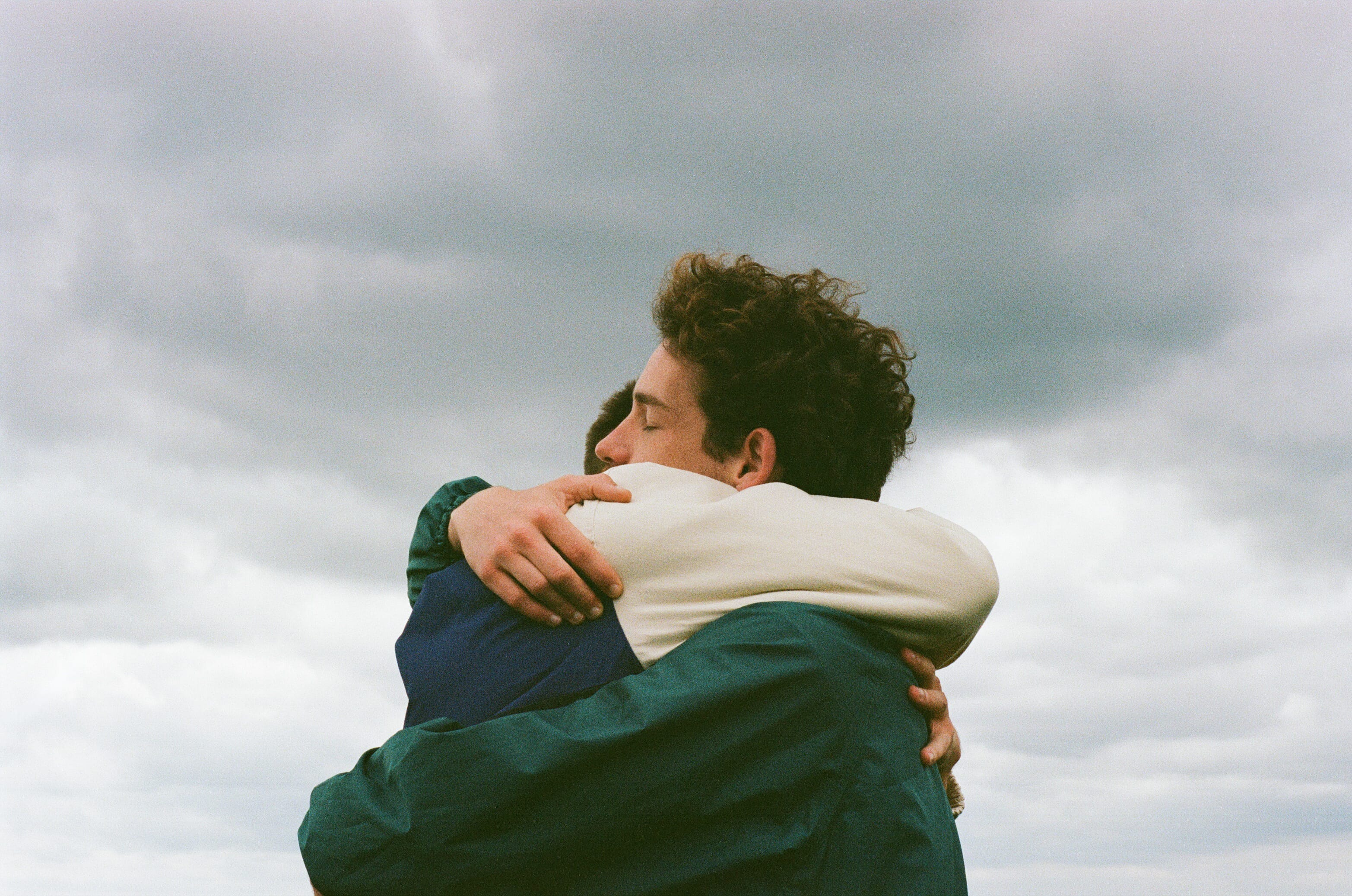 two men are embracing outdoors with a cloudy sky visible behind them
