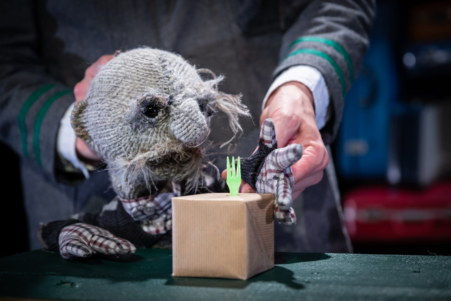 Small grey puppet looks at a green plastic fork.