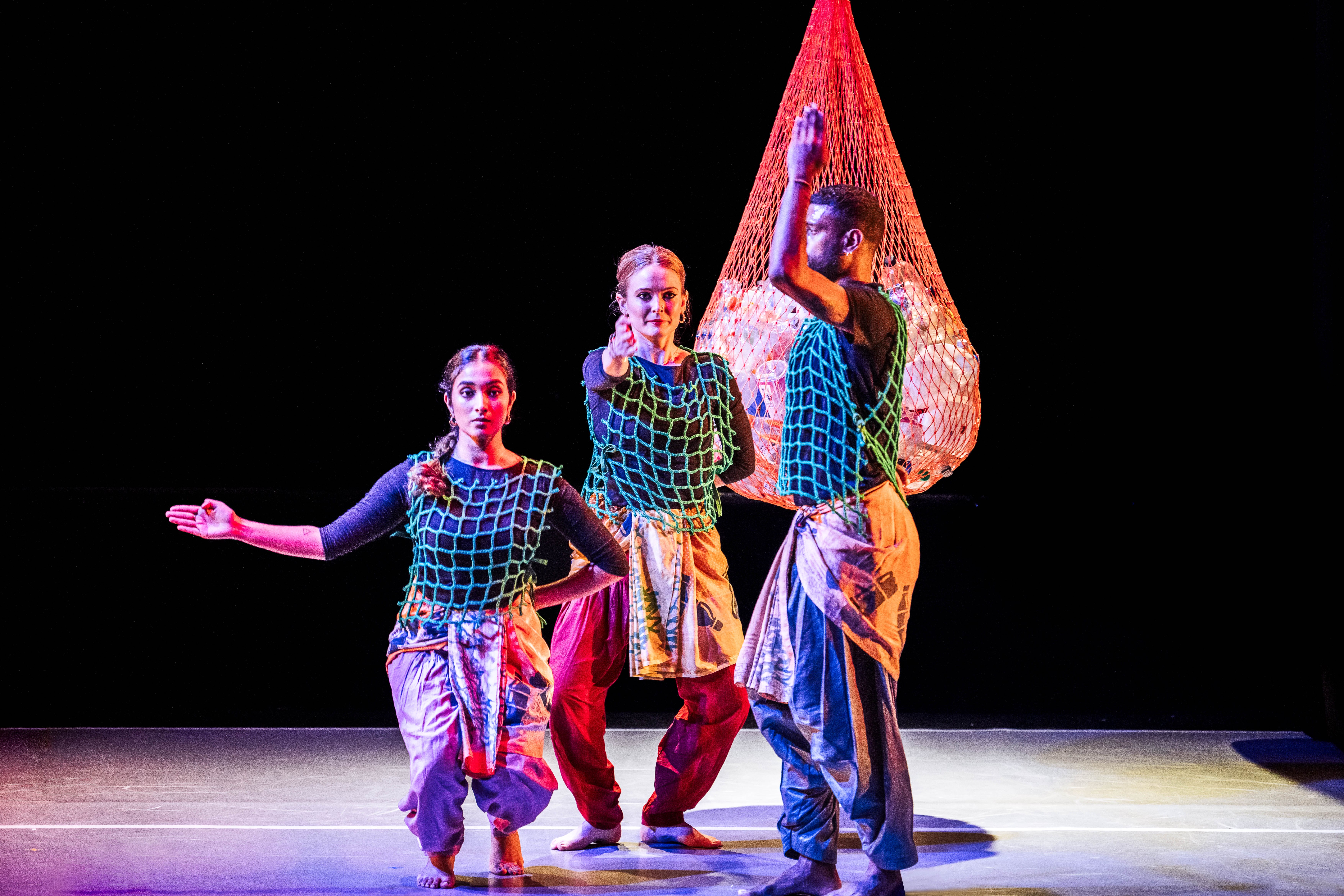 Three performers stand on stage, each standing in a different pose, wearing fishing net tops.