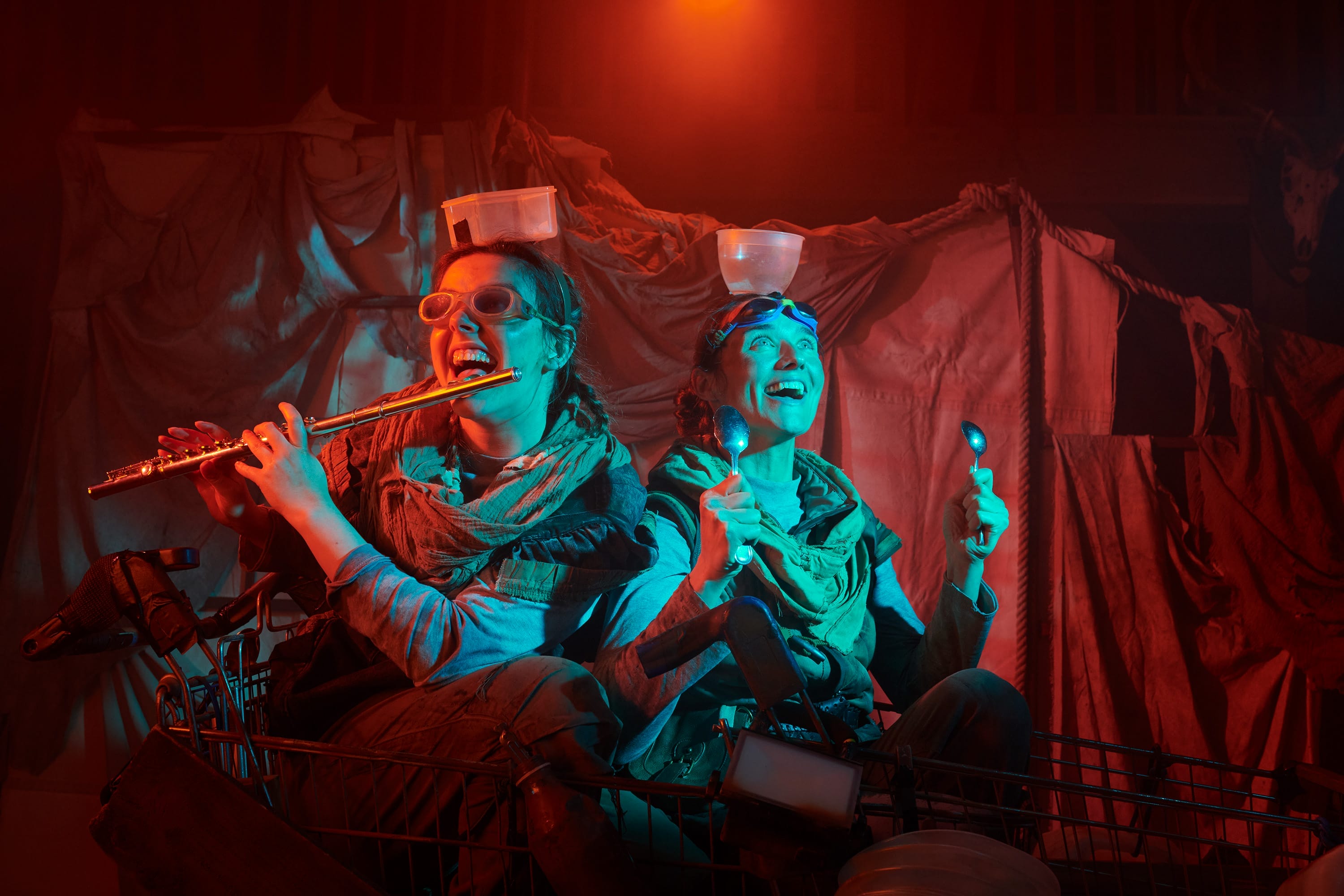 Two performers sit on stage with plastic bowls balanced on their head, one playing the flute.
