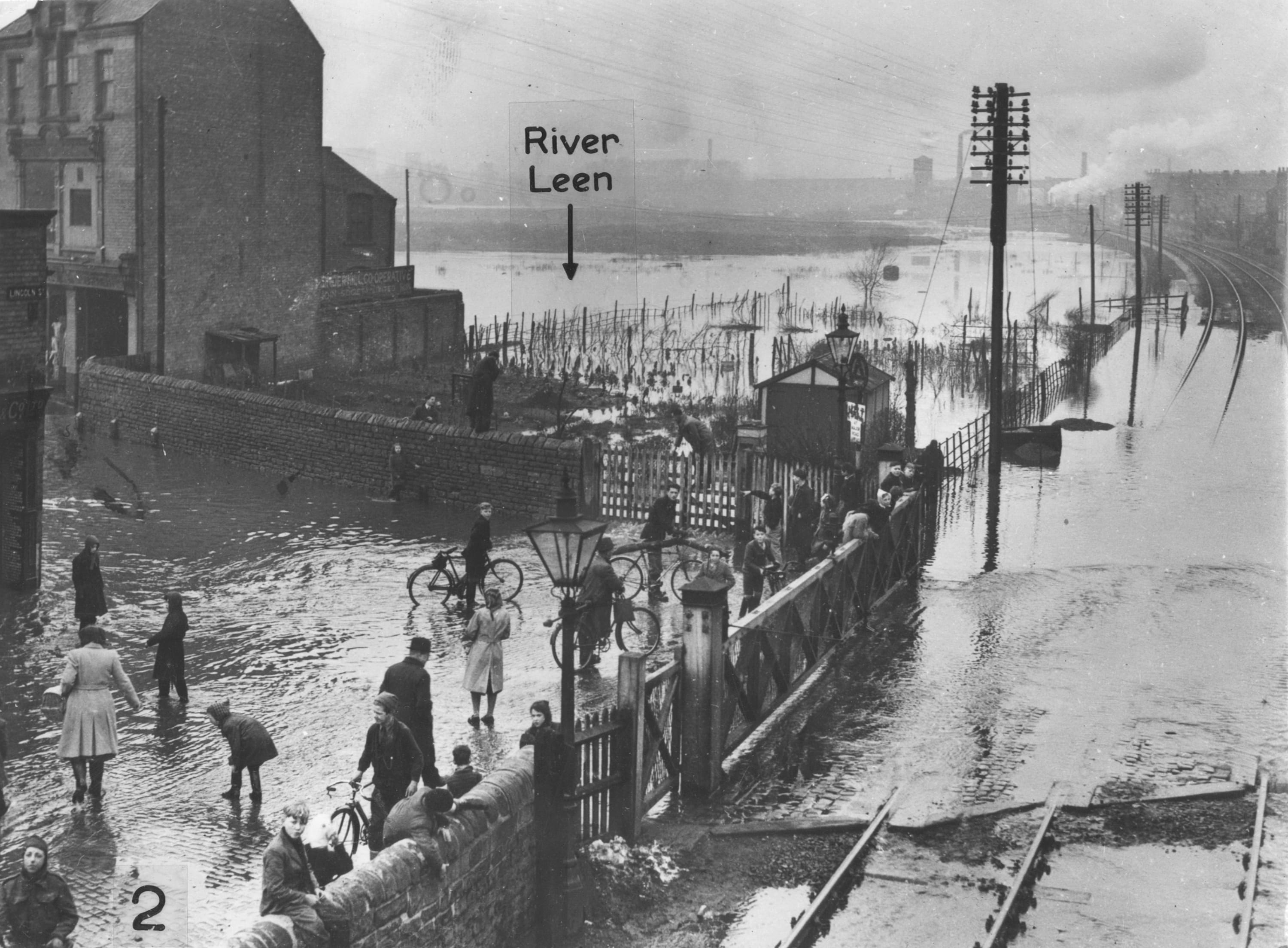 Photograph of flooded Basford Crossings, March 1947, showing the flooded railway tracks and road; pedestrians and cyclists wait at the railway gate end of David Lane. The view has been taken facing northwards, possibly from railway footbridge, and annotated to show the course of the River Leen.
