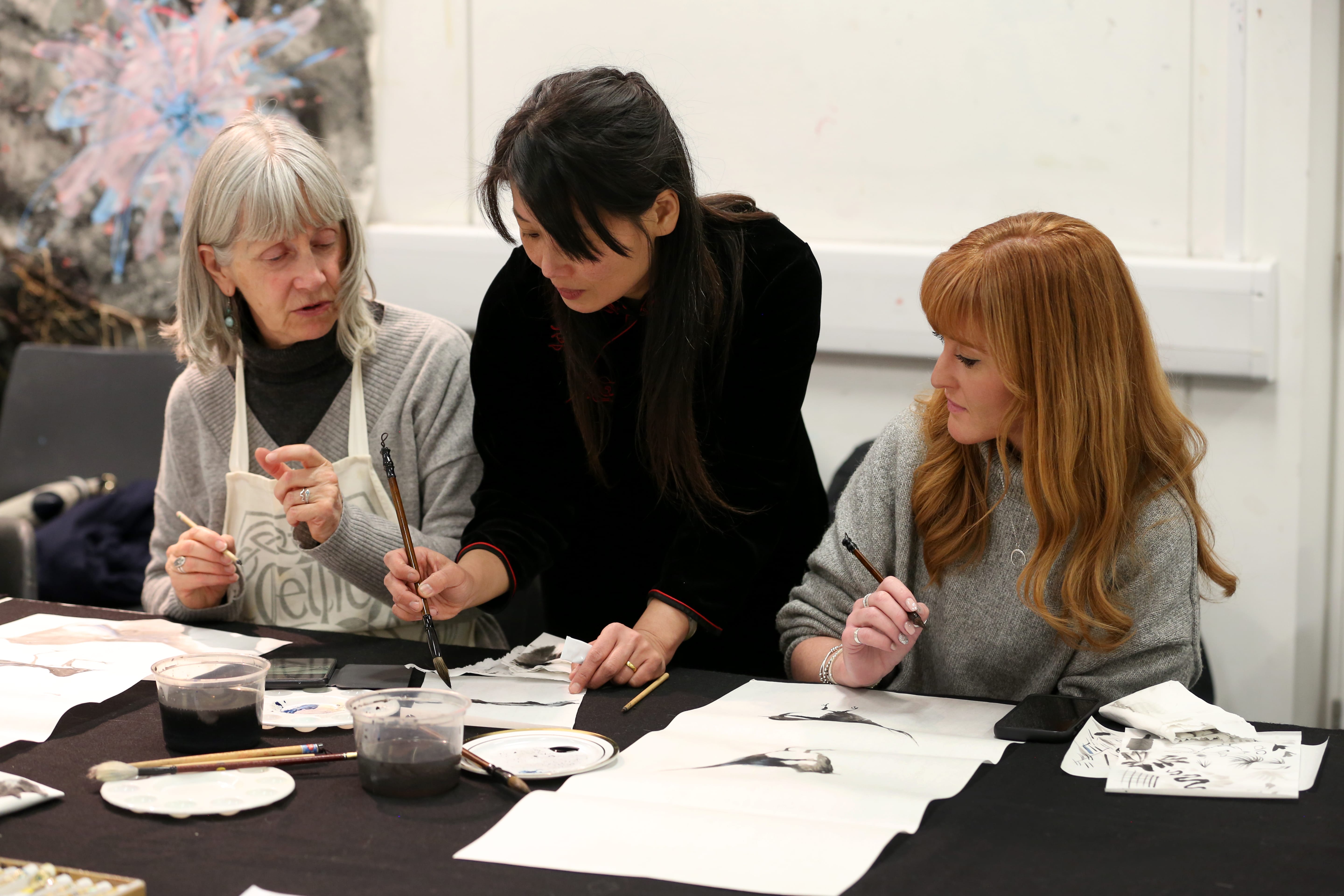 The image shows the instructor bent over the table helping two women with their brushwork