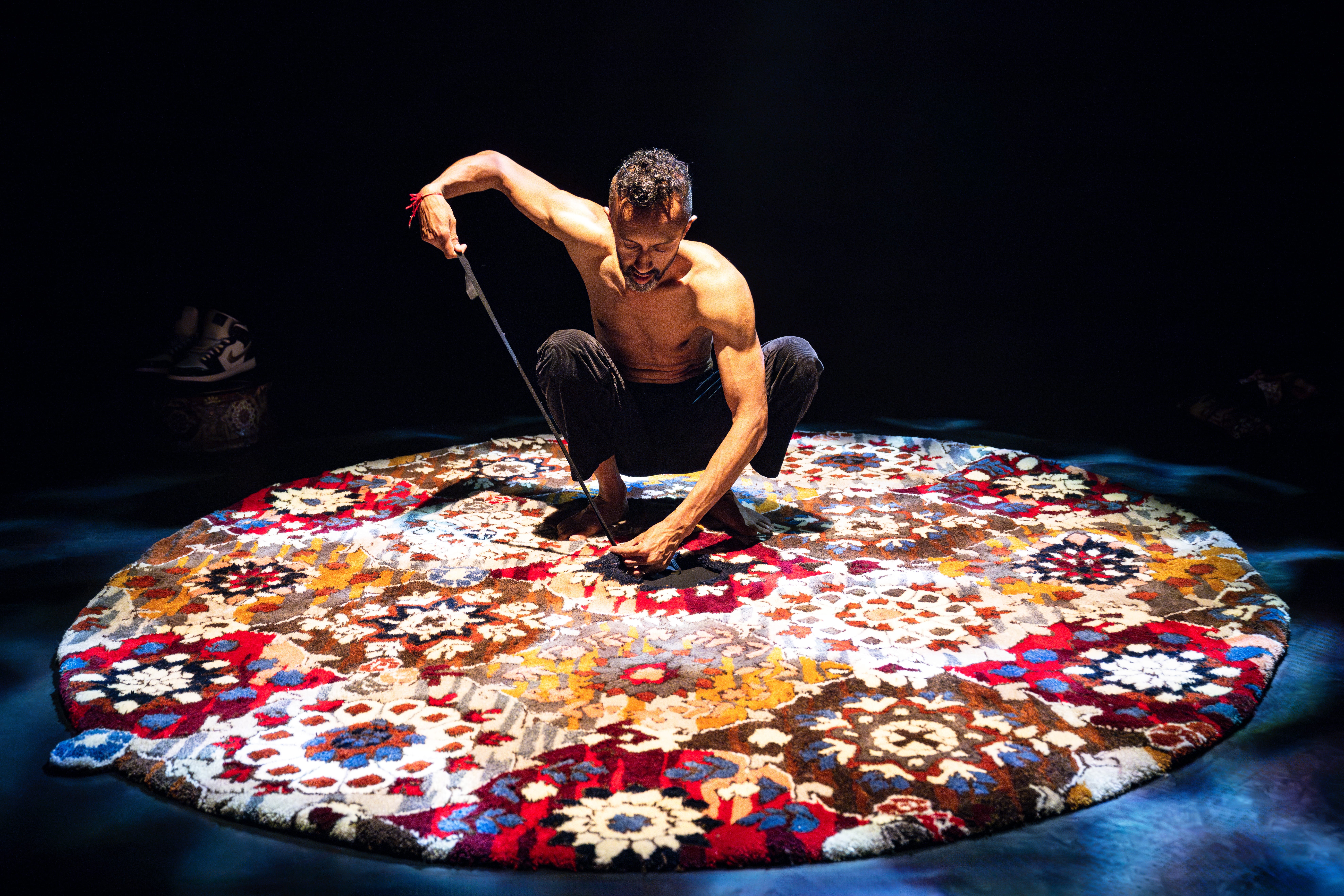 Hetain crouches down on a colourful carpet on a dark stage