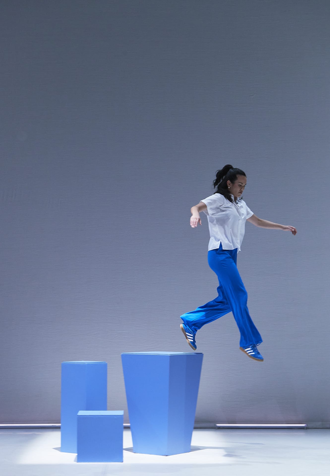 A dancer wearing blur trousers and a white shirt is in mid air jumping off a blue box.