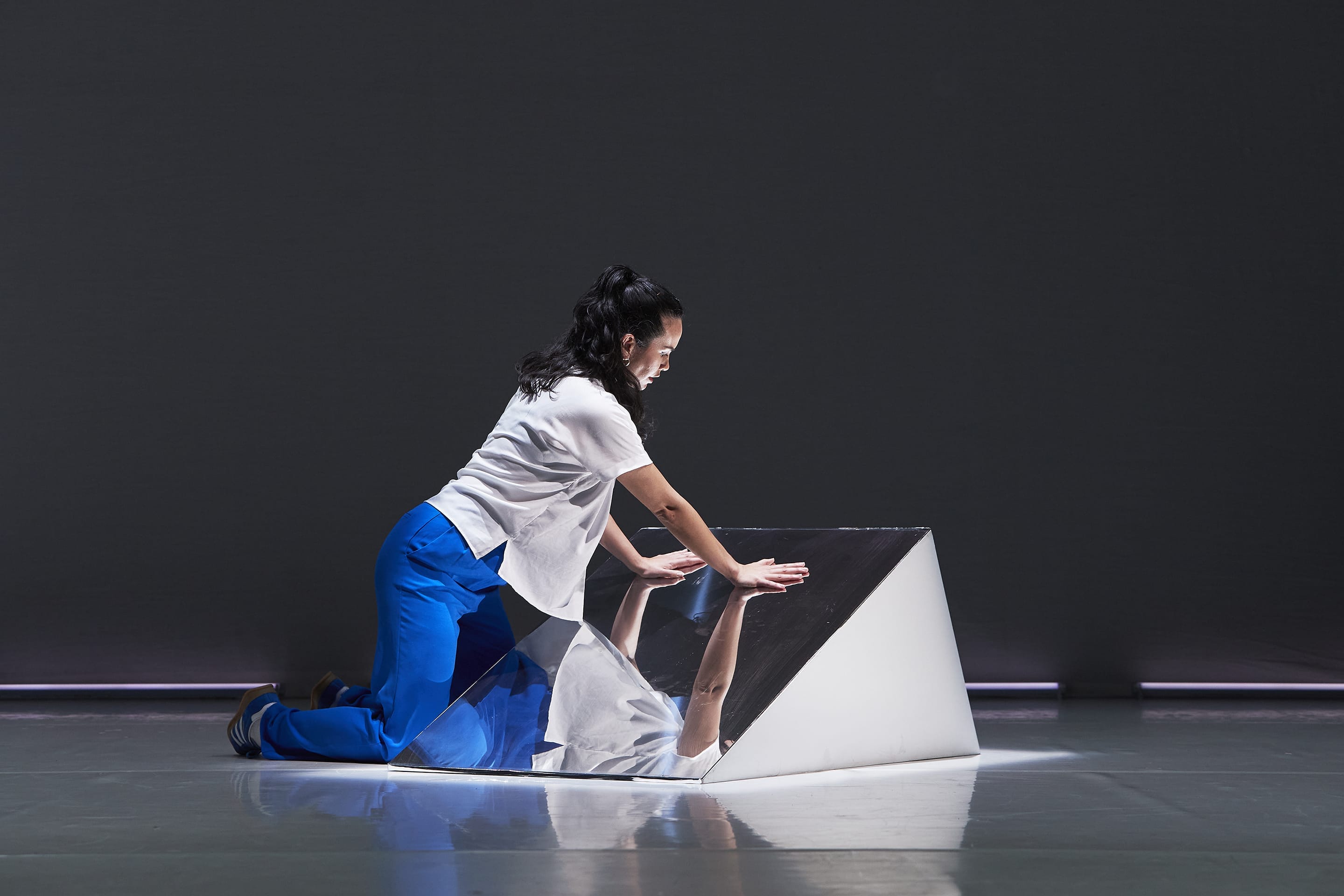 A woman wearing blue trousers and a white shirt is kneeling next to a raised mirror on a stage.