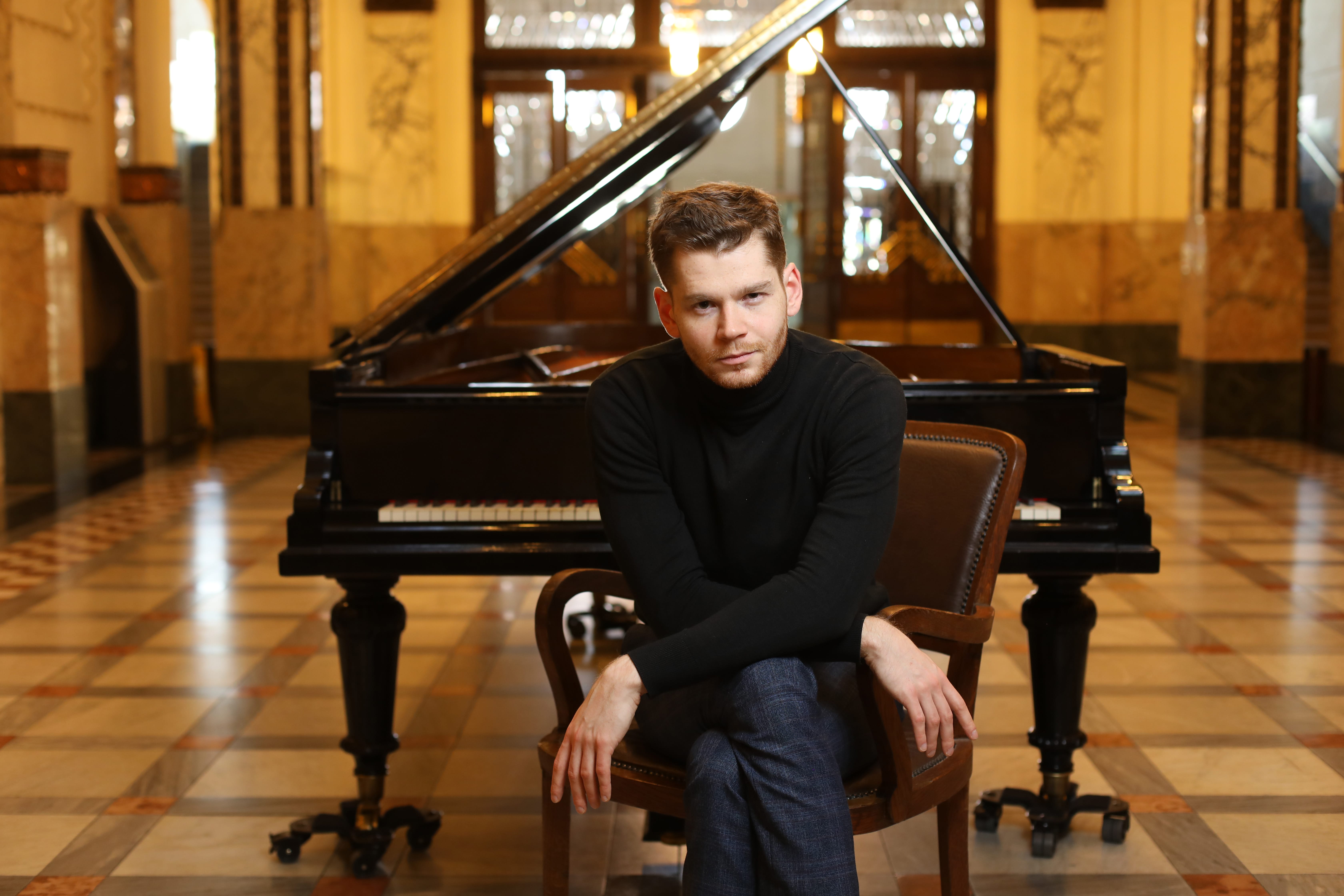 Andrey Gugnin is sat in an opulent room in front of a grand piano, leaning towards the camera.
