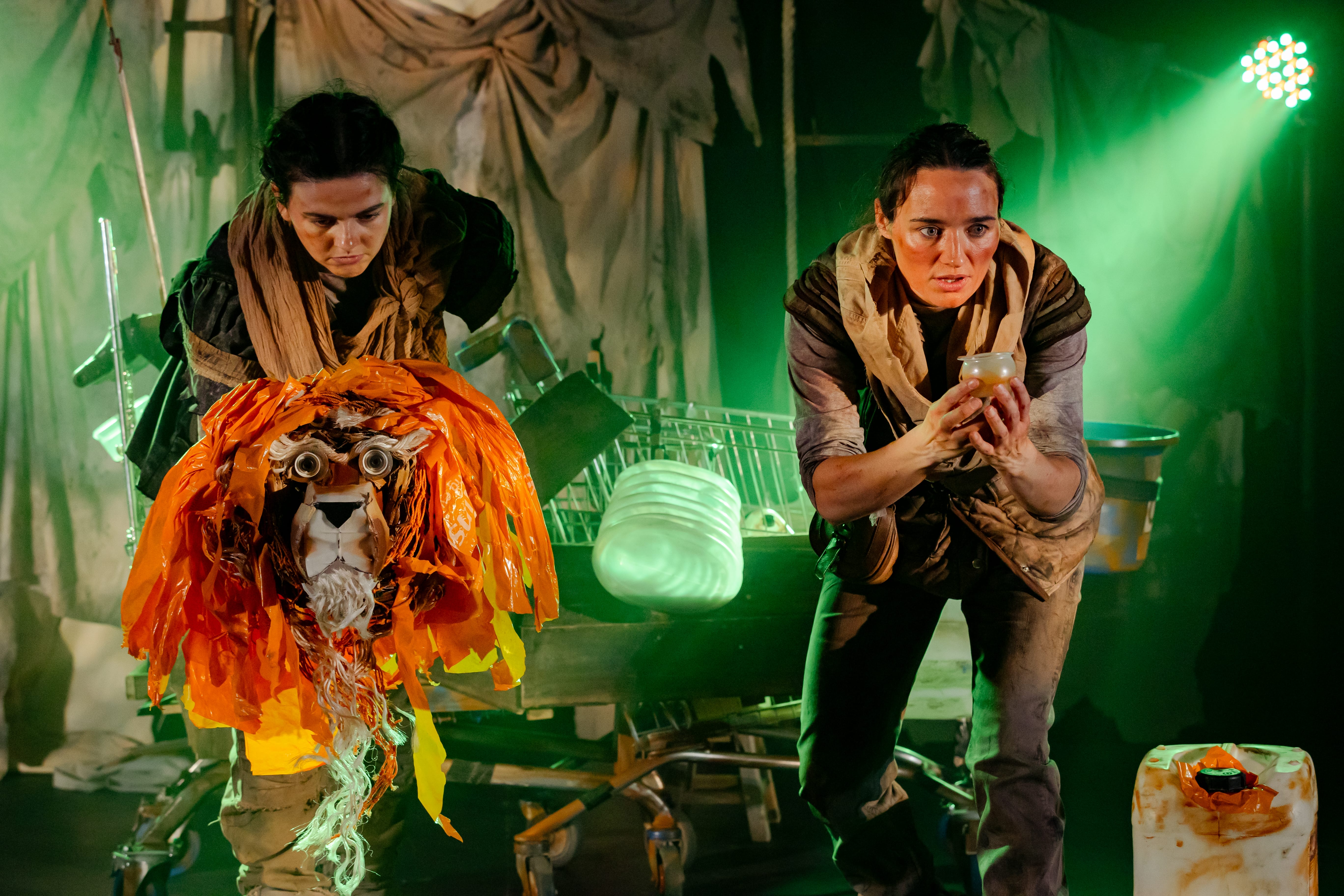 two actors on stage holding plastic objects and a lion head made of plastic scrap