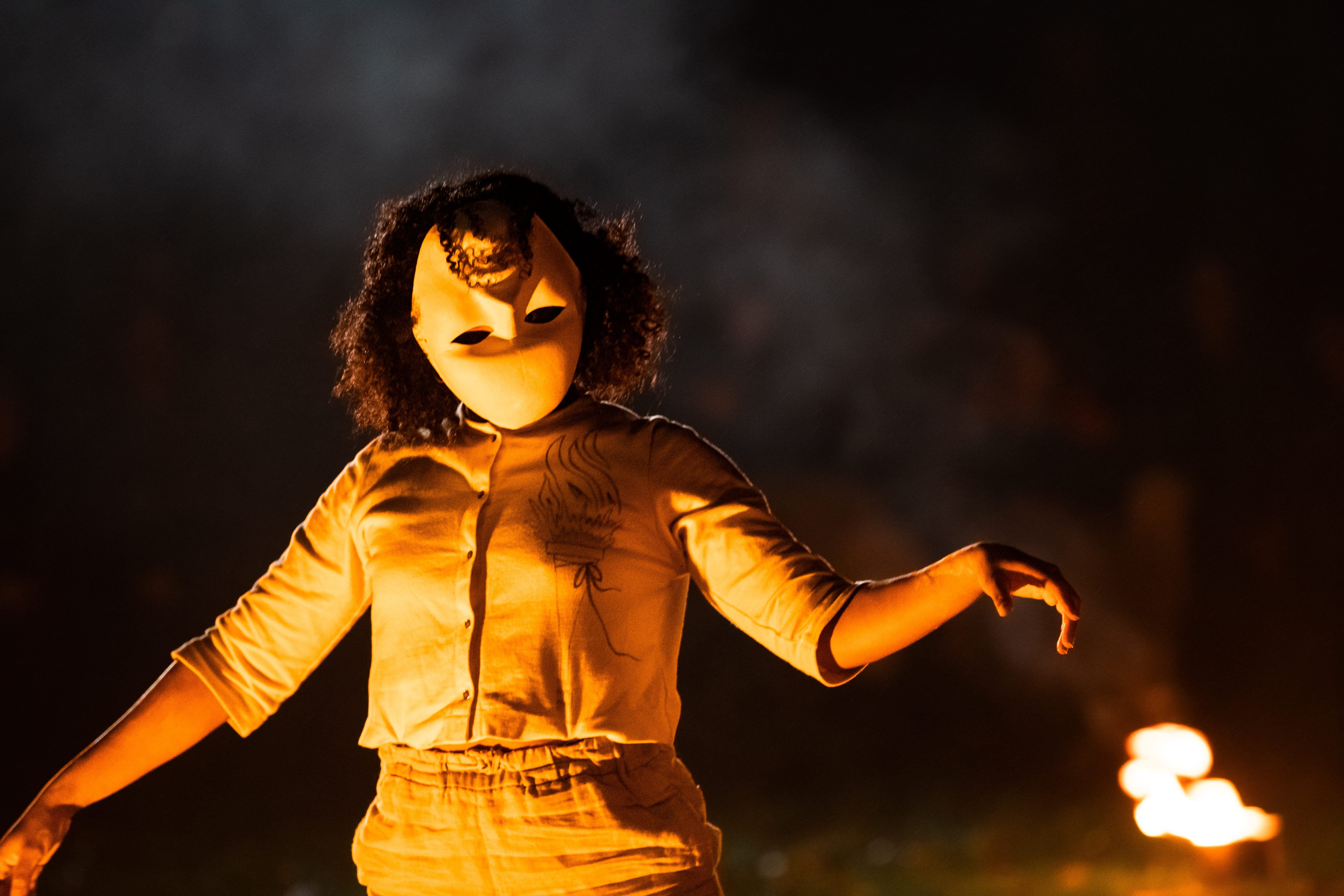 an actor dressed in white wearing a mask upside down, with the light of a fire falling on her