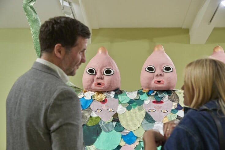 Two visitors of the exhibition are in the foreground while song dynasty ooo remains in sharp focus in the background, two heads of the costume staring back at the camera