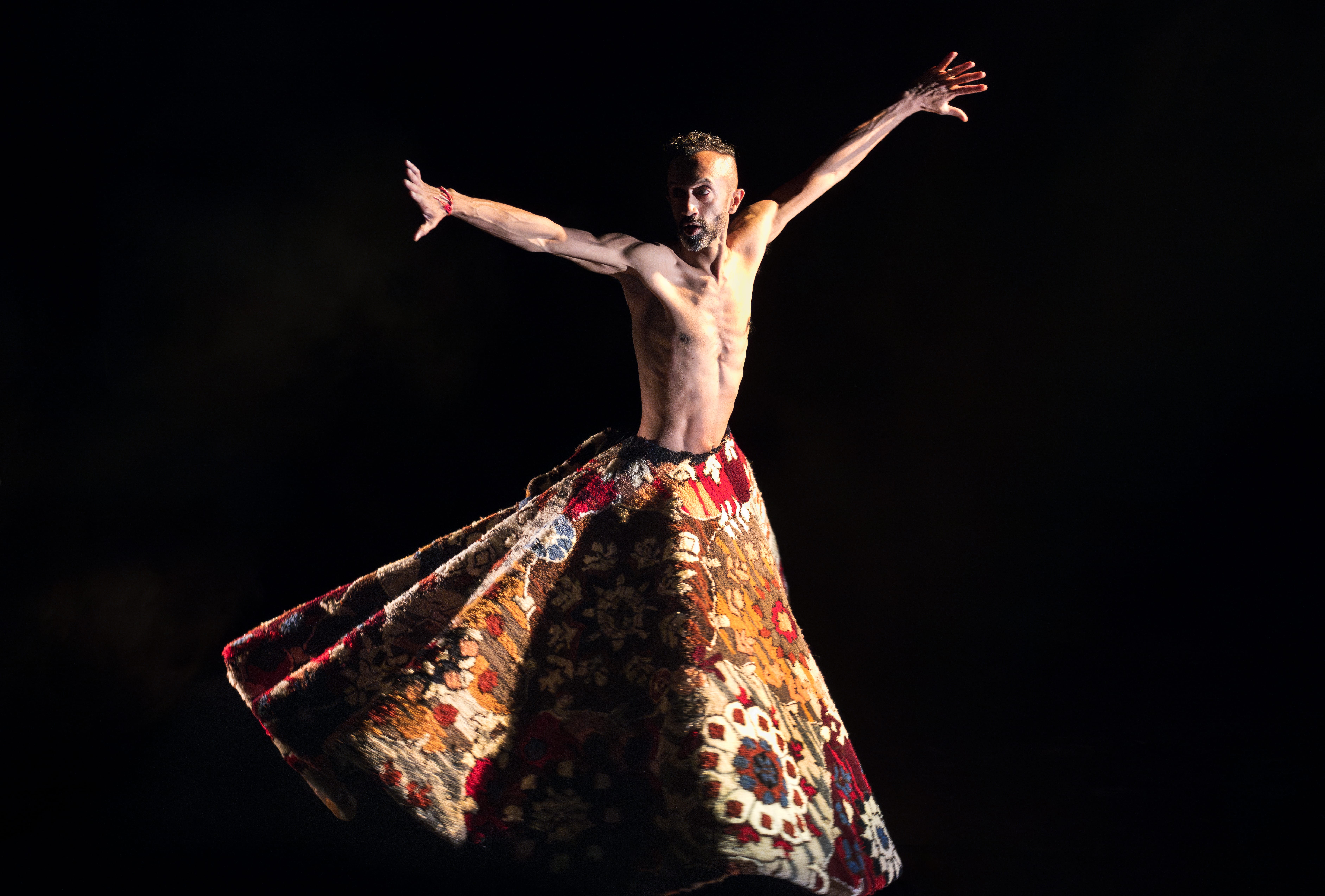 Hetain Patel dances illuminated on a dark, shadowy stage wearing an ornate costume that flows out dramatically