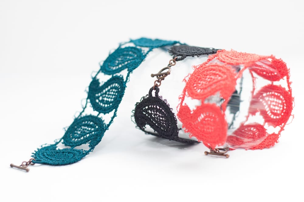 Three bracelets made of lace in a paisley design. They are teal, black and a pinkish orange.