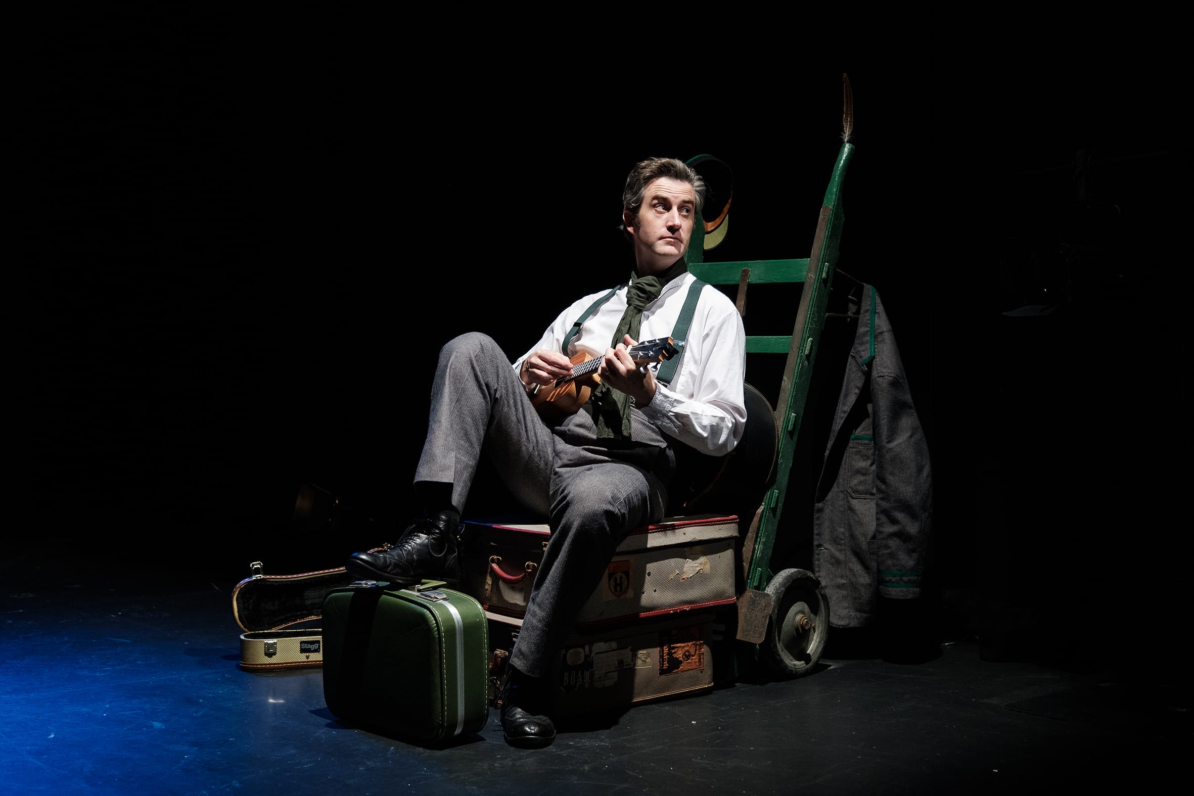 A performer holding a guitar sits on a luggage trolley