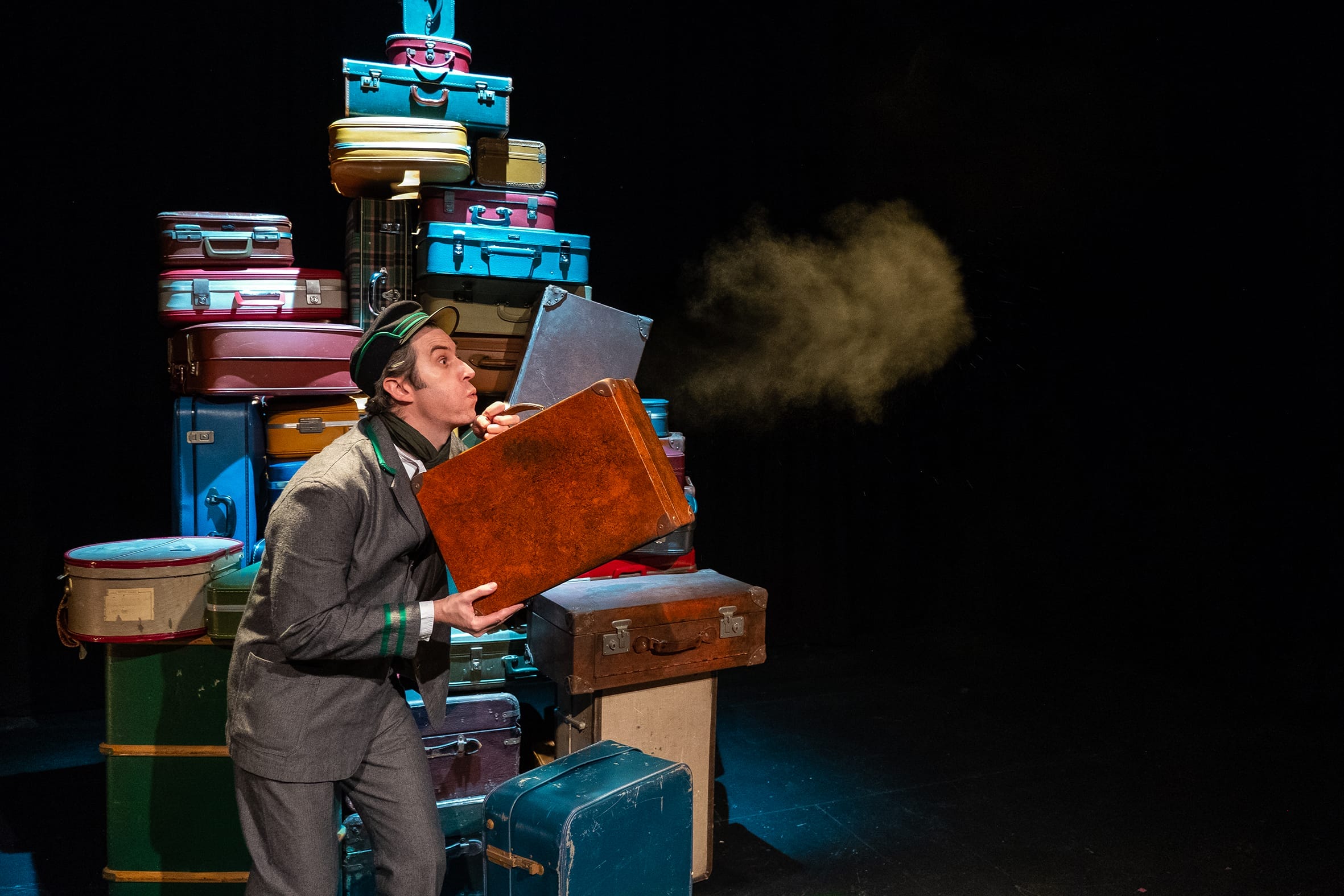 A performer stands on stage with a pile of suitcases behind