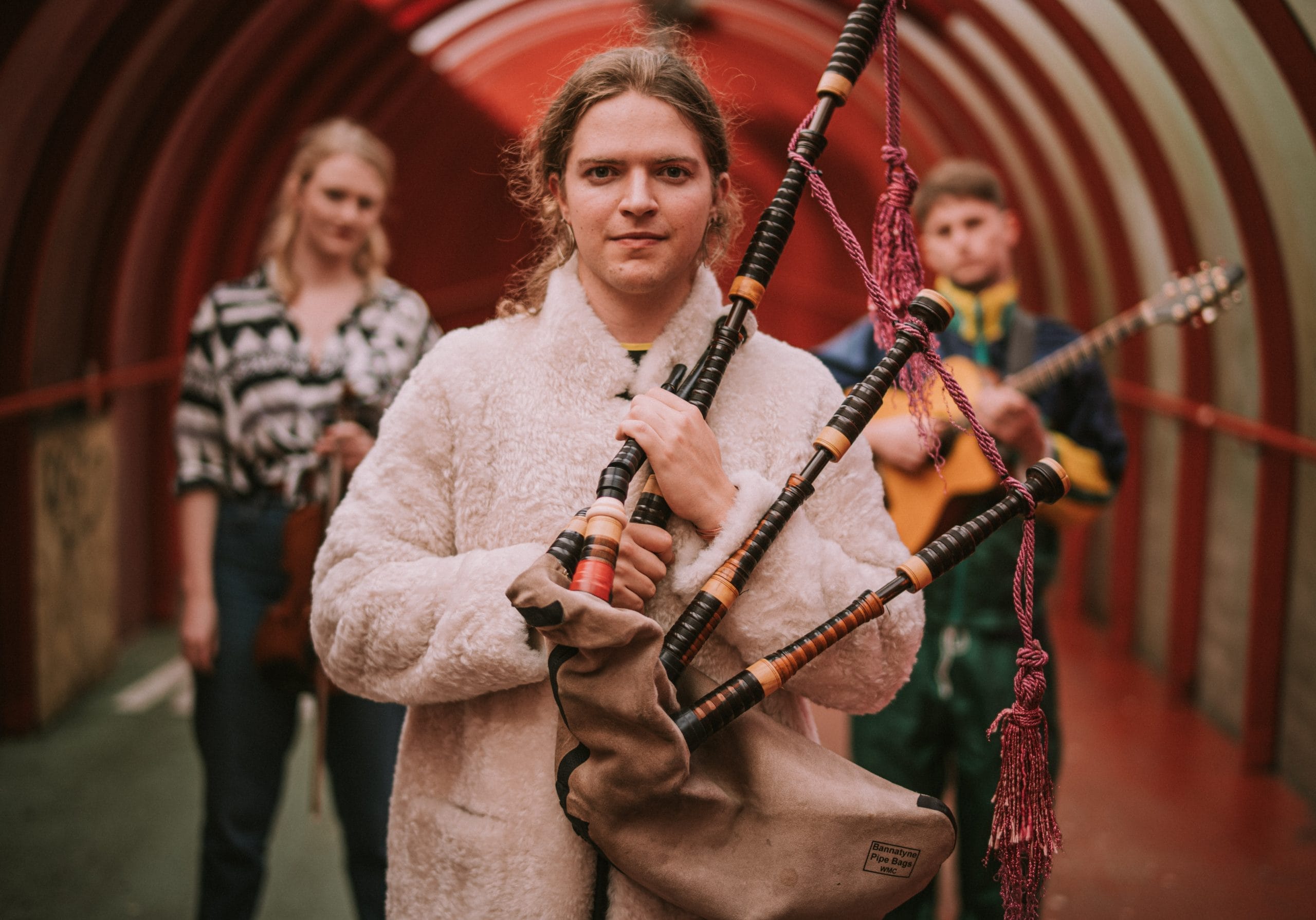 Malin Lewis stands in the forefront with their two-octave bagpipe while the other musicians, Sally Simpson and Ali Hutton are a few steps back in the background.