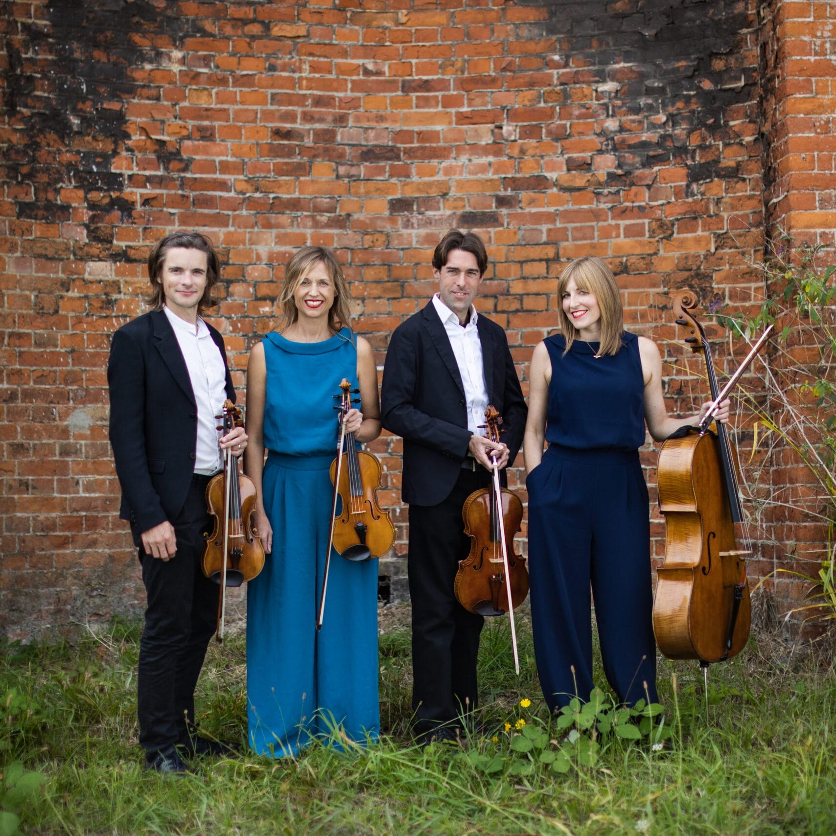 The quartet stand smiling with their instruments against a brick wall