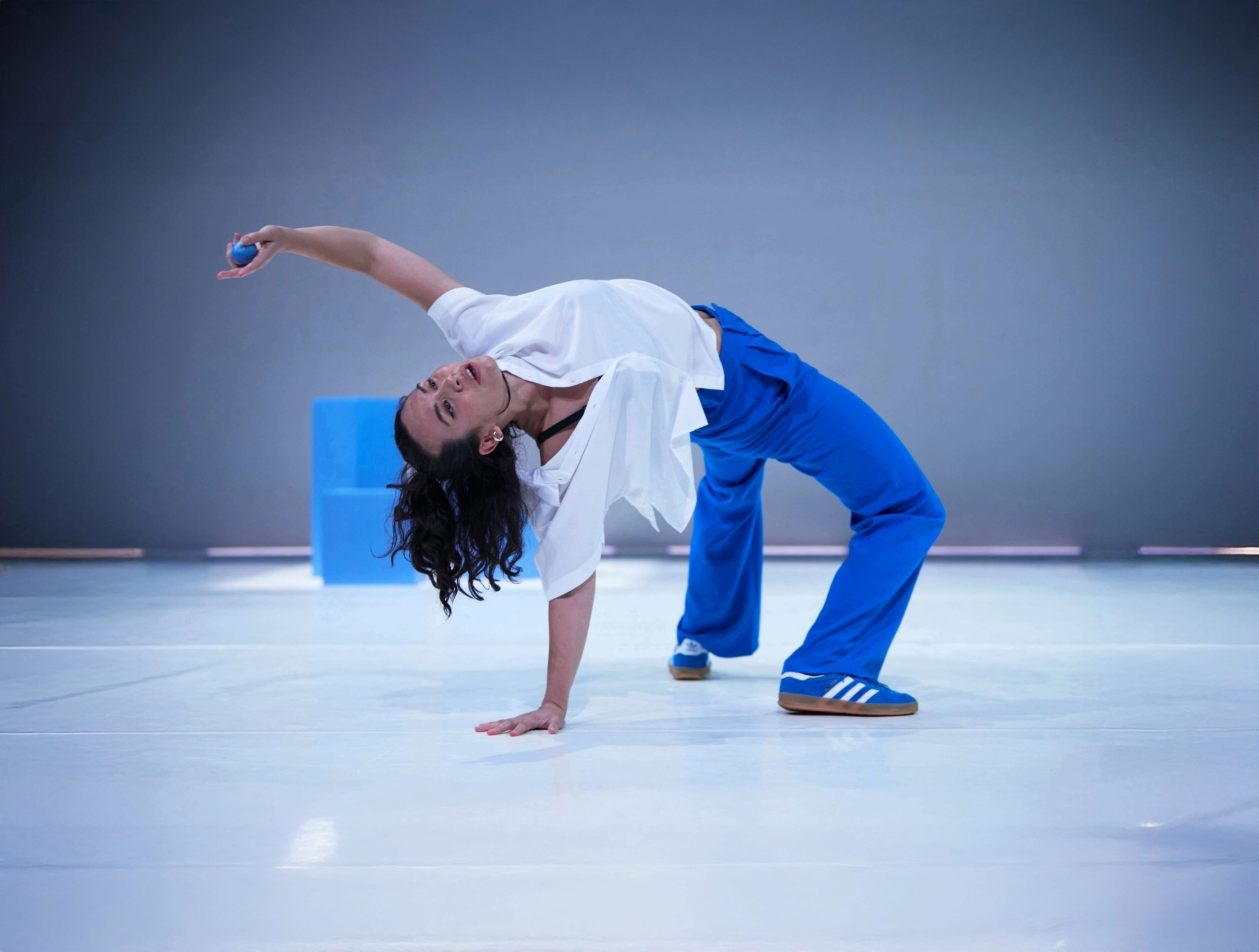 A dancer wearing blue trousers and a white shirt takes up a crab position in a white studio space.