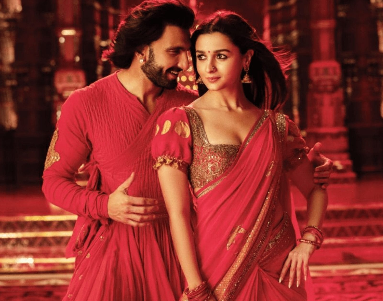 A joyful couple, a man and a woman, are embracing each other in a grand room of red and gold. The man, on the left, has dark hair and a beard, and is looking towards the woman, he is wearing a red cross over top with gold embellishment. The woman, on the right, has dark, wavy hair and is looking across the man with a subtle expression. She is wearing a colourful, intricately patterned top and a sari. Both appear to be in their 30s.