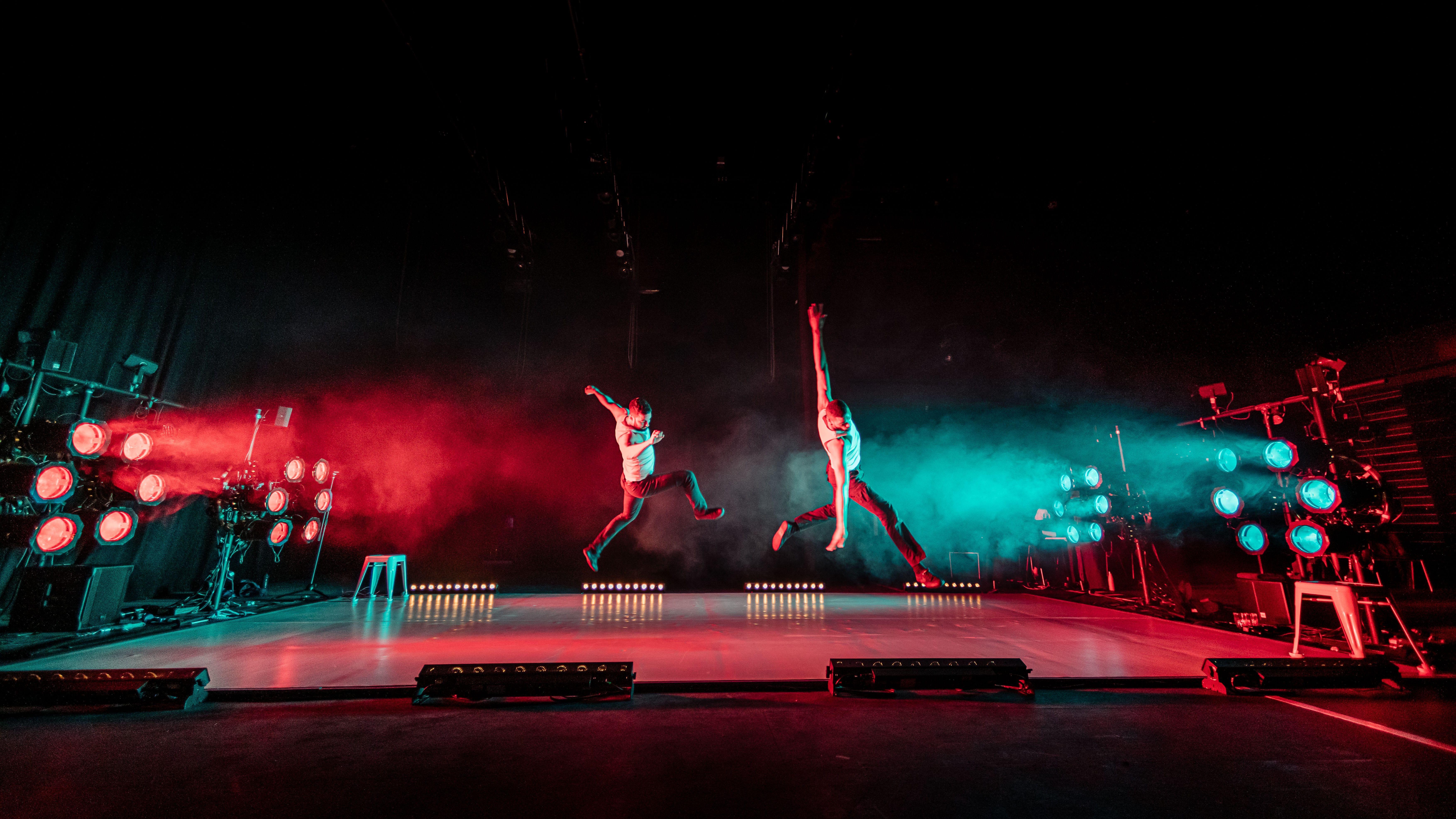 Two performers leap across stage, back lit with blue and red lights.