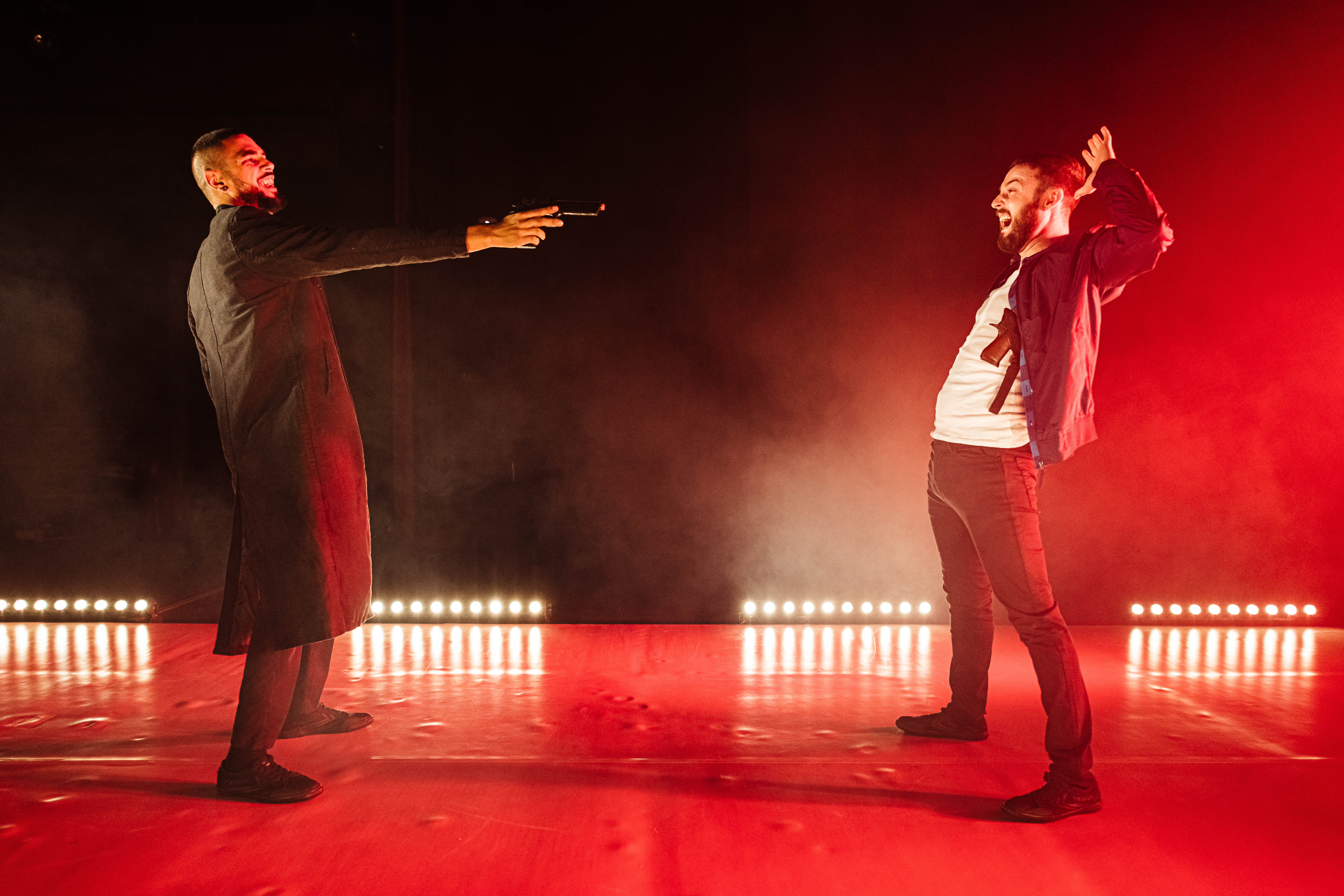 Two men on a red stage perform. One of them has a mock gun pointed to the other who has both his hands up in the air