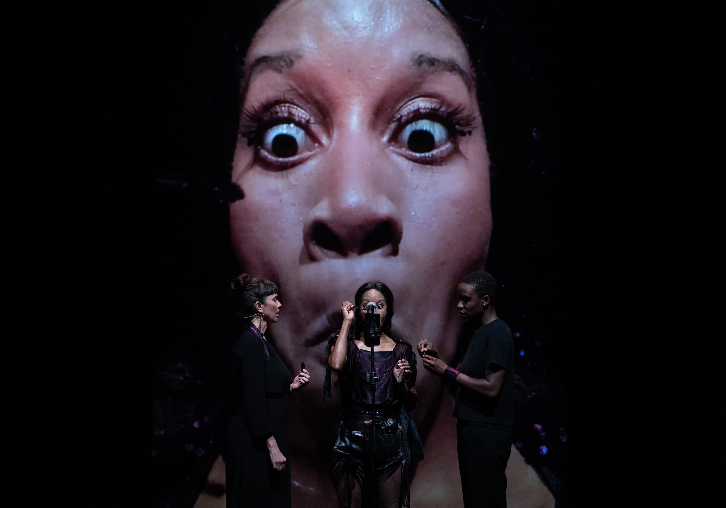 Three people performs on stage while one of their faces is projected in big behind them