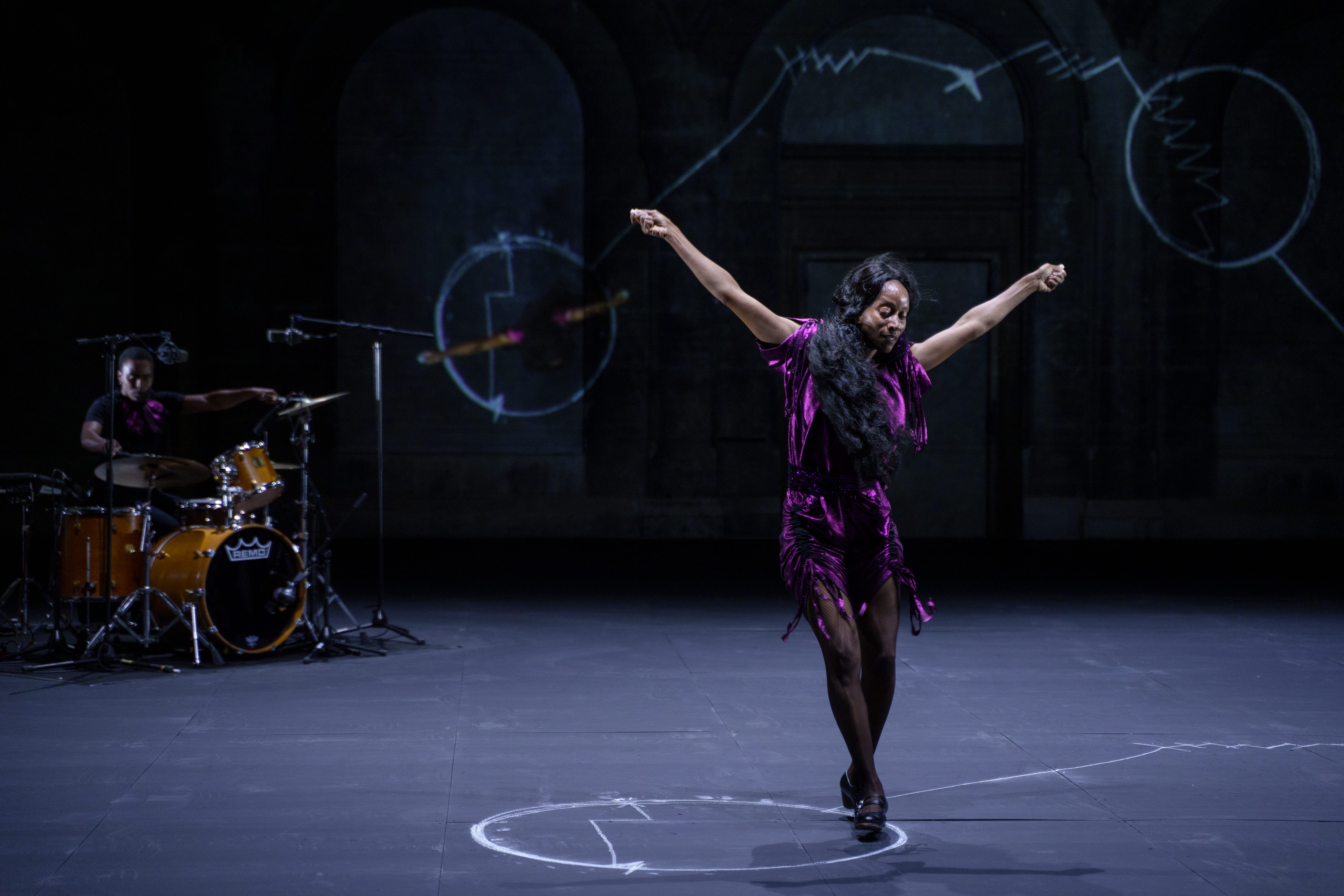 A woman performs flamenco on a dark stage with a musician in the background