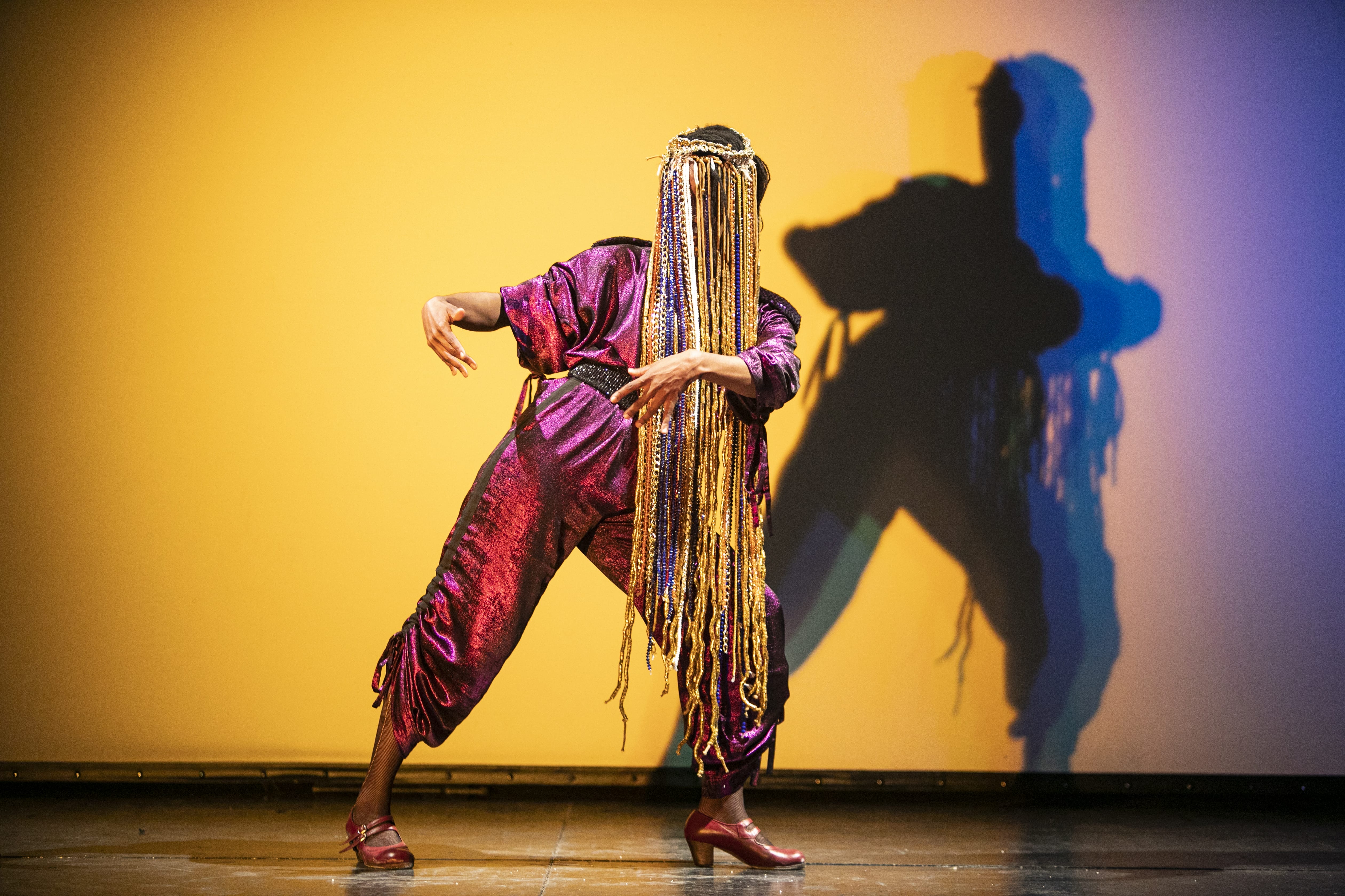 A dancer stands on stage, wearing red, with braids that nearly reach the floor, in front of a yellow background.