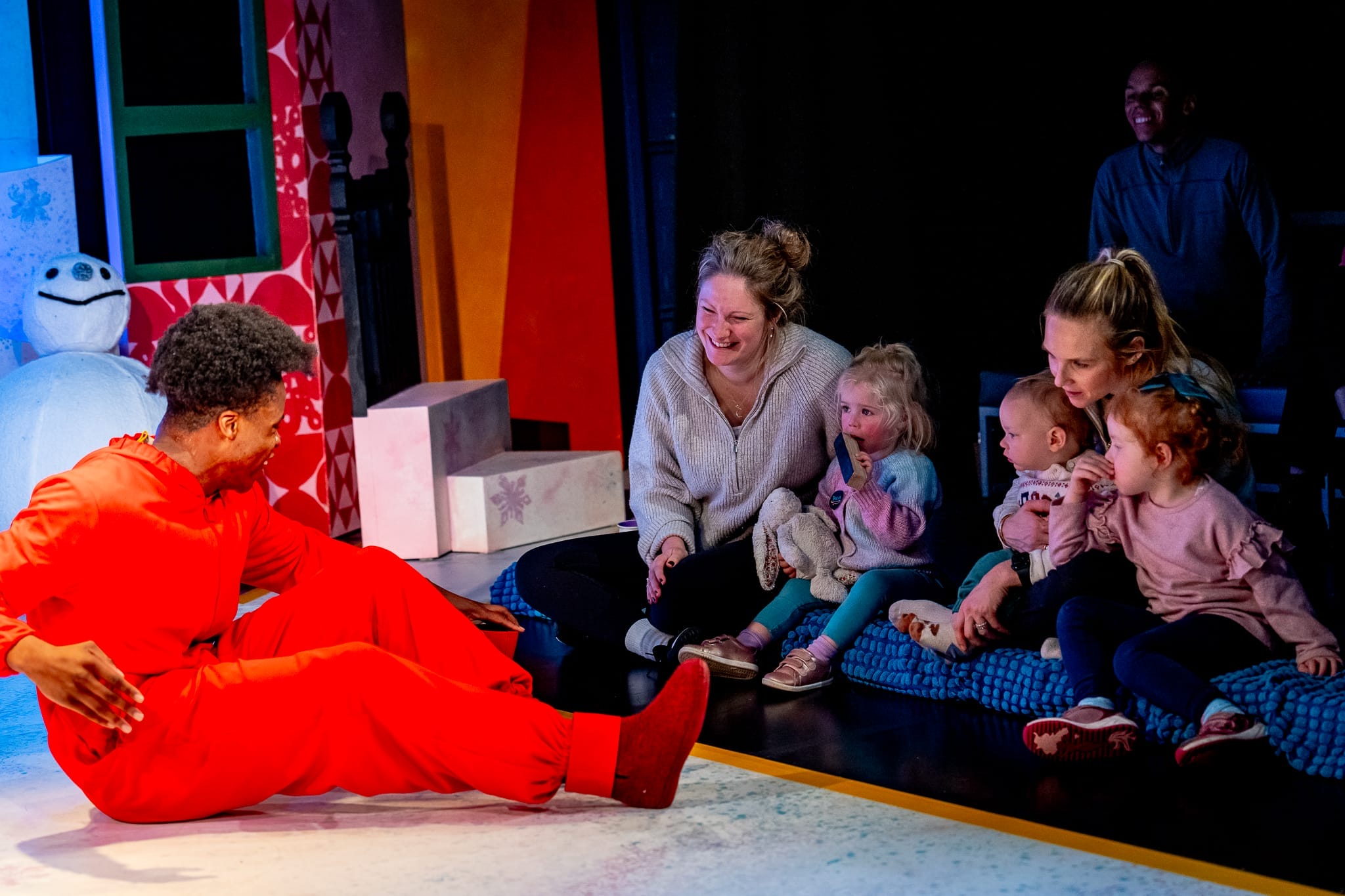 An actor sits on the stage and talks to a group of children and their parents sitting alongside the edge of the stage