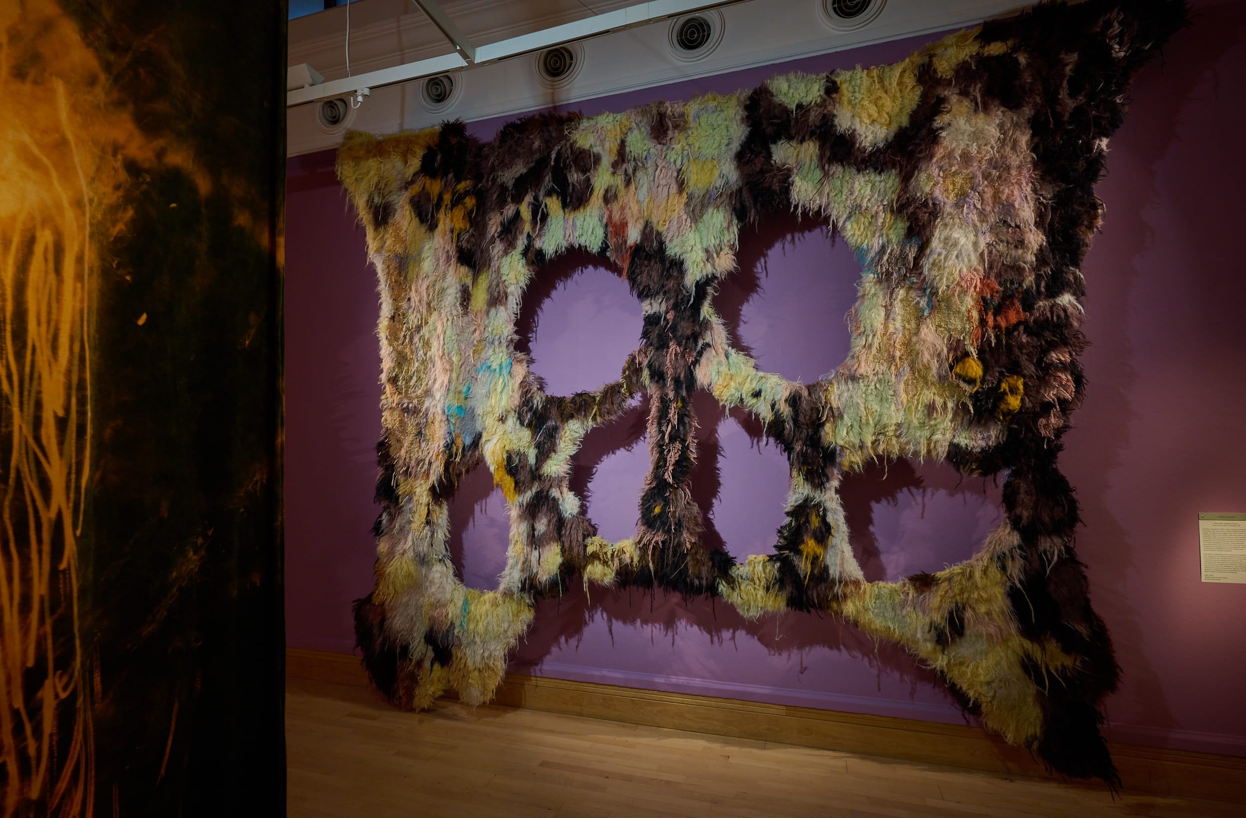 Picture shows Achaintre's piece; it is a giant textile hanging from a steel rod, it ressembles a primitive, savage, yet colourful mask or bemoaning creature.