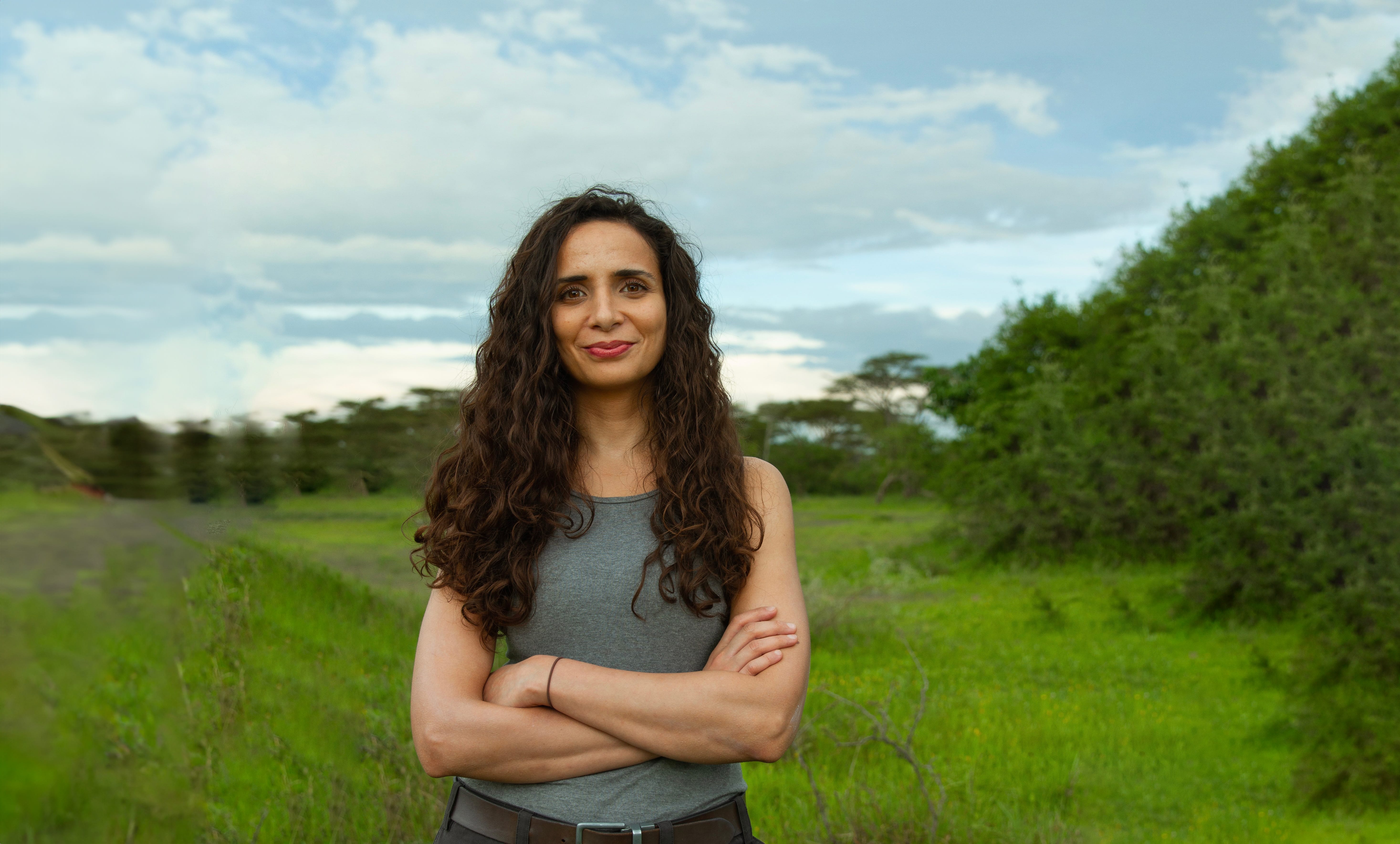 A smiling woman with long dark hair stands with her arms crossed in the countryside