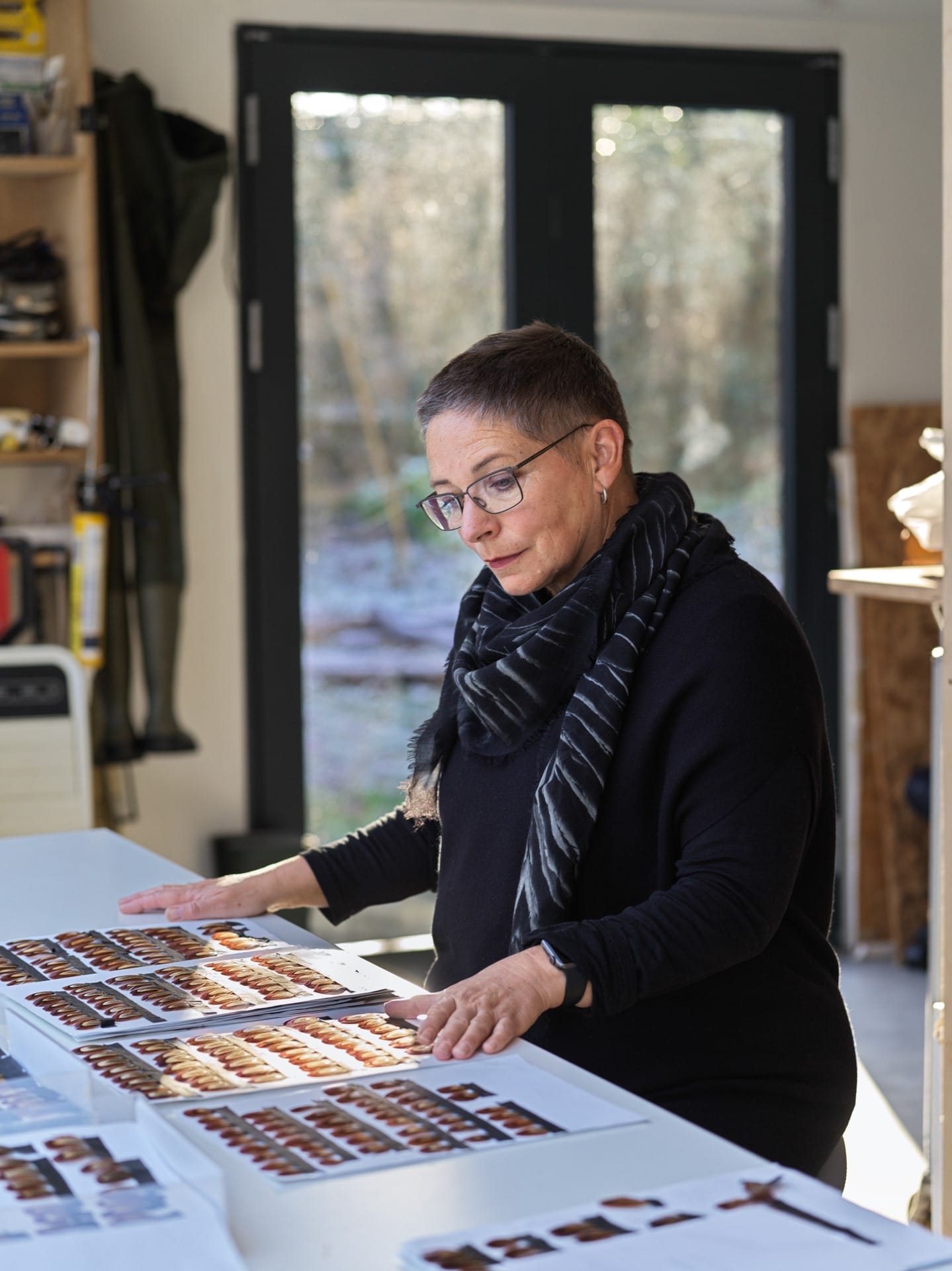 Artist Kate MccGwire is stood looking at various feathers laid out on a work surface in front of her