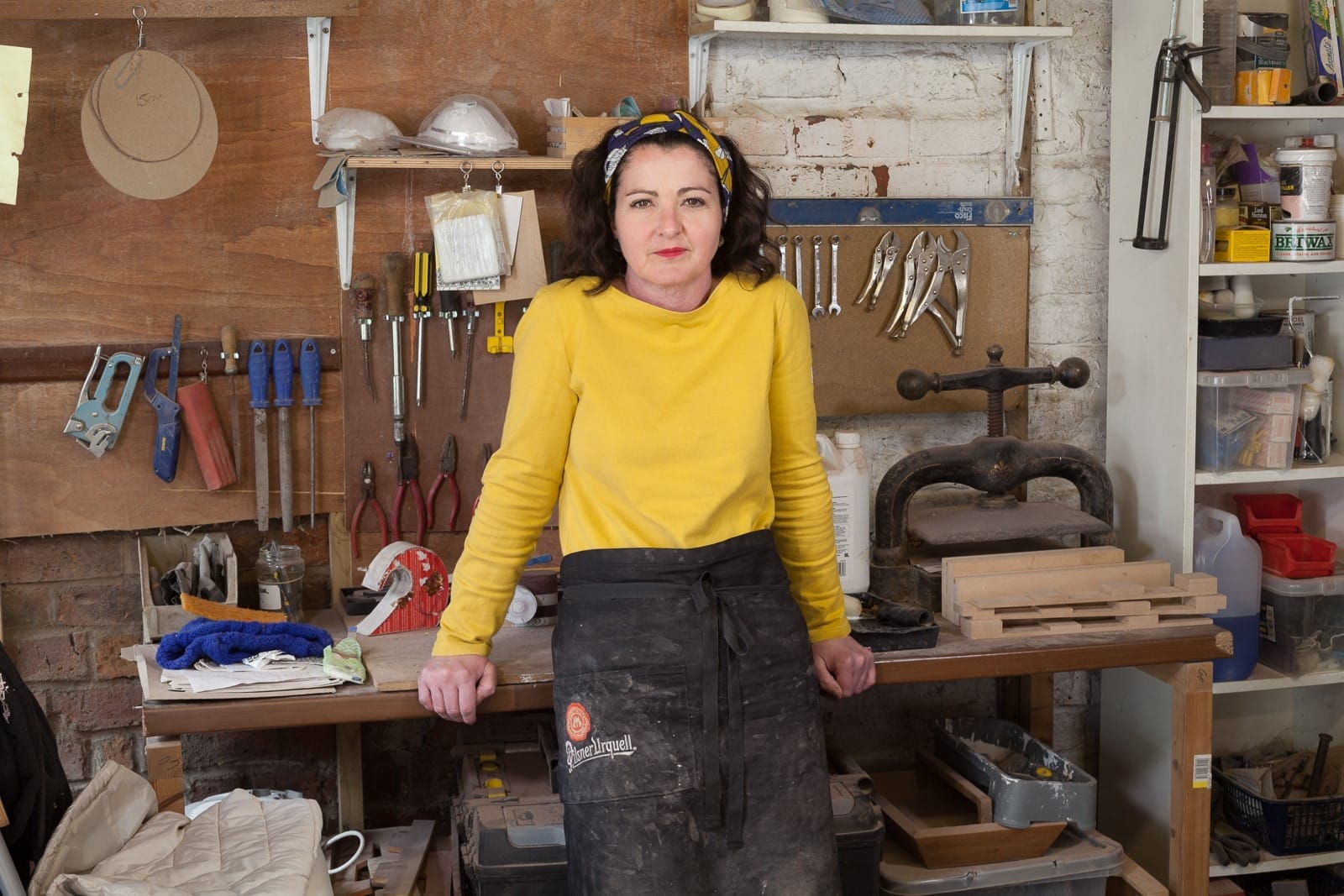 Artist Hannah Lane in her workshop. She is wearing a yellow jumper and leaning on her workbench