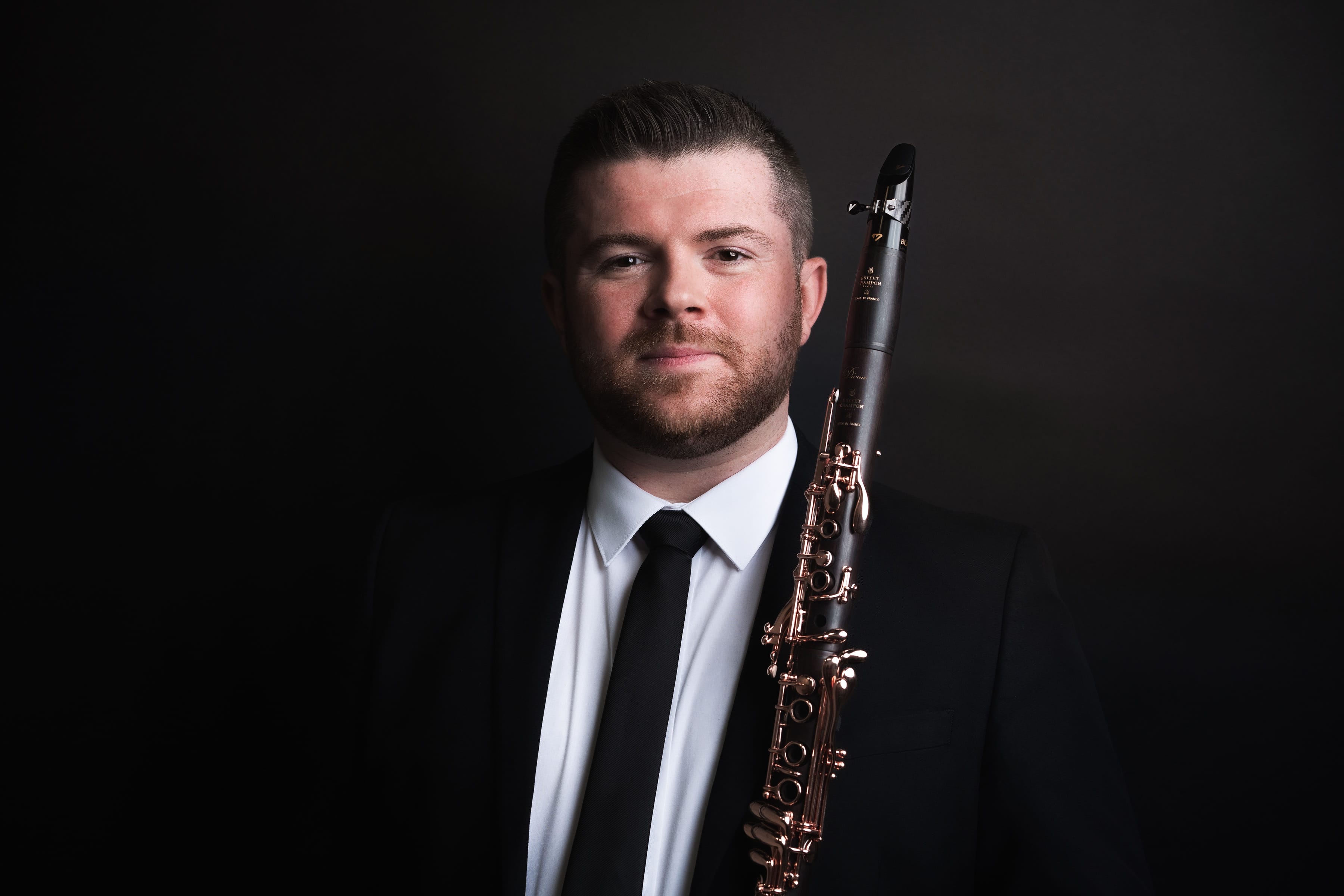 Musician Julian Bliss stands, wearing a black suit and tie, with his clarinet.