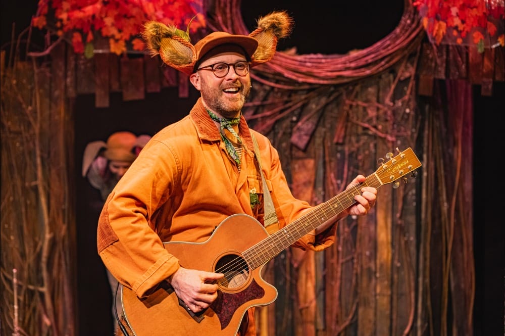 A man plays the guitar, wearing an orange coat and a hot with fluffy ears.