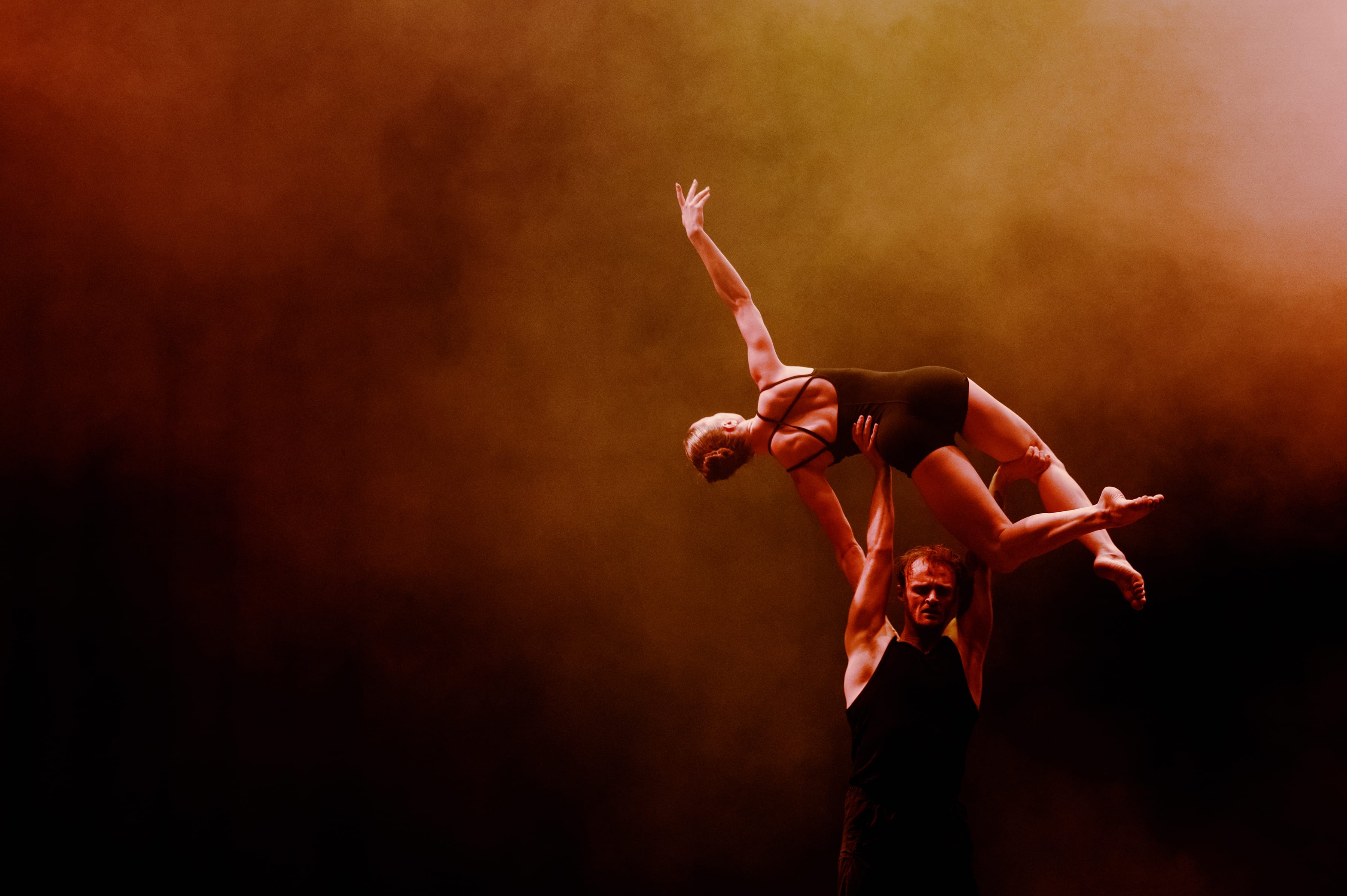 A male dancer lifts a female dancer, wearing a black leotard, above his head, with low lighting and haze in the background.
