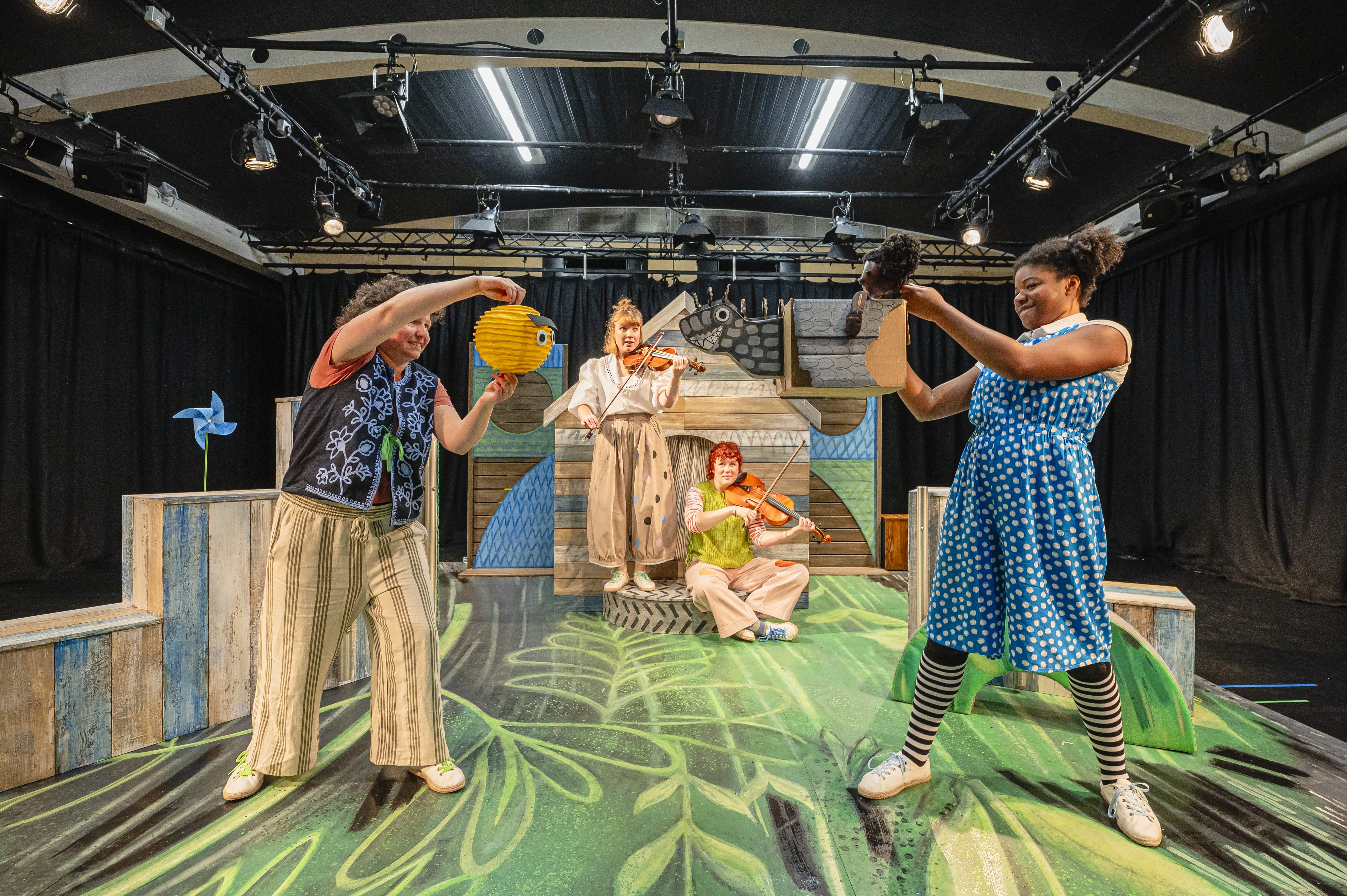 A stage is set, with a green floor - two people at the back play violins and two actors stand at the front with puppets.