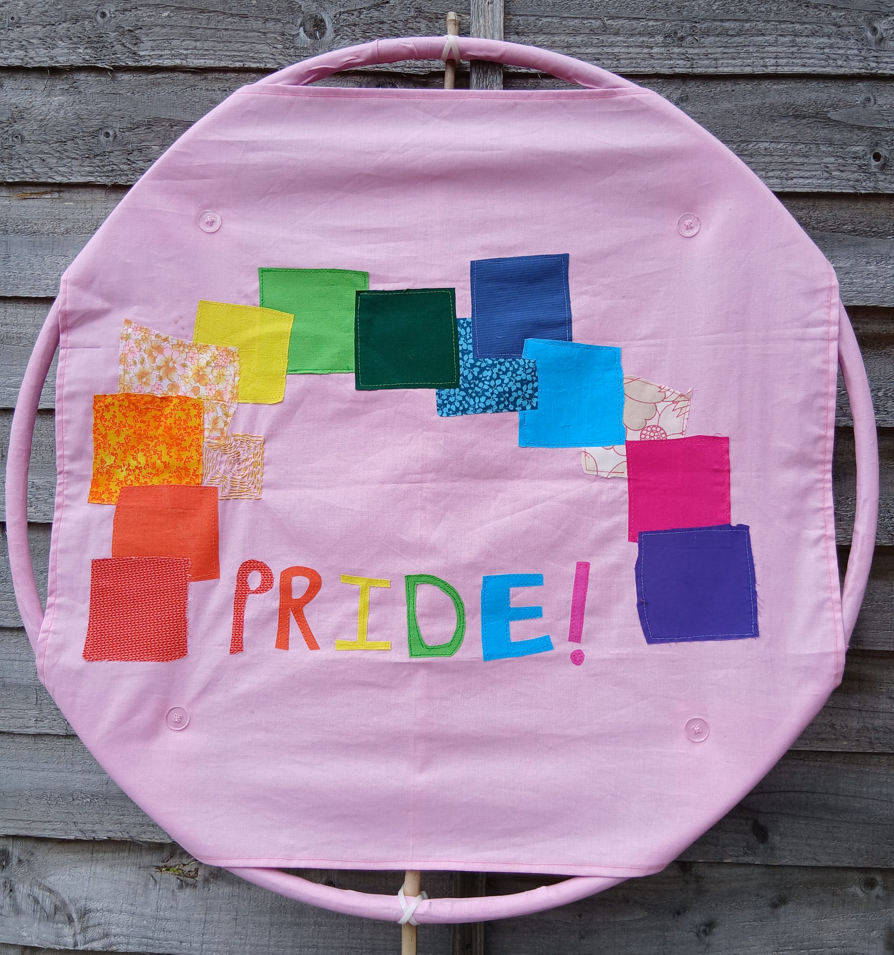 a photo of a pale pink fabric on a round frame with squared shaped colourful fabrics and the letters forming the word PRIDE! added on with applique method