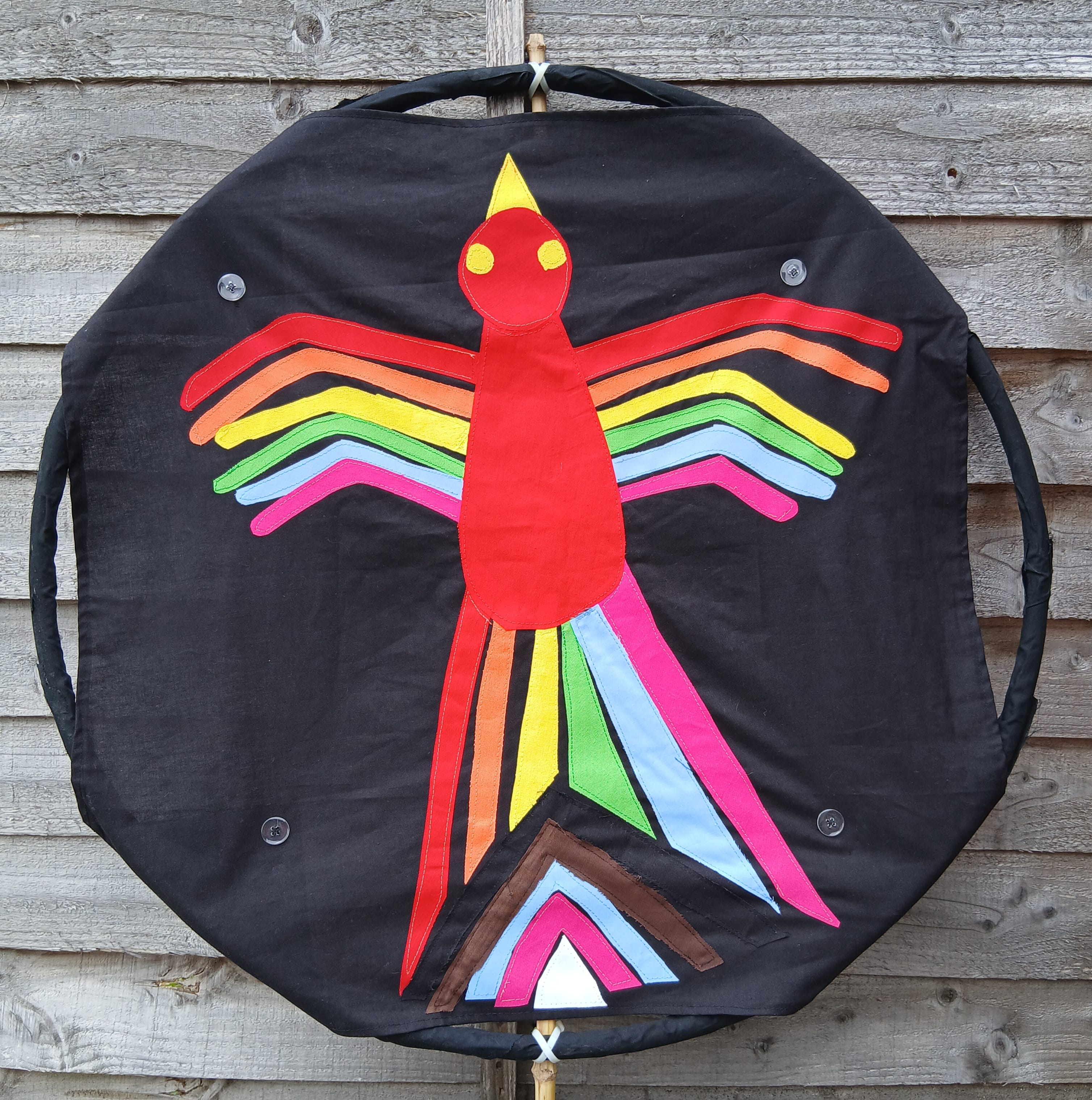 a photo of a black fabric on a round frame with variously shaped colourful fabrics added on to form a flying bird with applique method