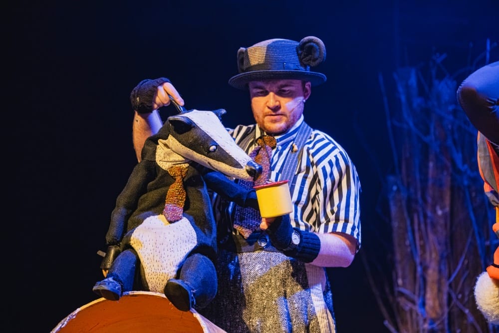 A man stands on stage, wearing a black and white striped shirt and braces, stands with a puppet of a badger as it drinks from a cup of tea.