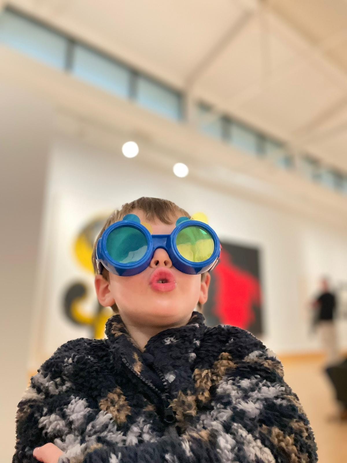 A small boy, wearing round glasses with colourful lenses looks at artwork in wonder.