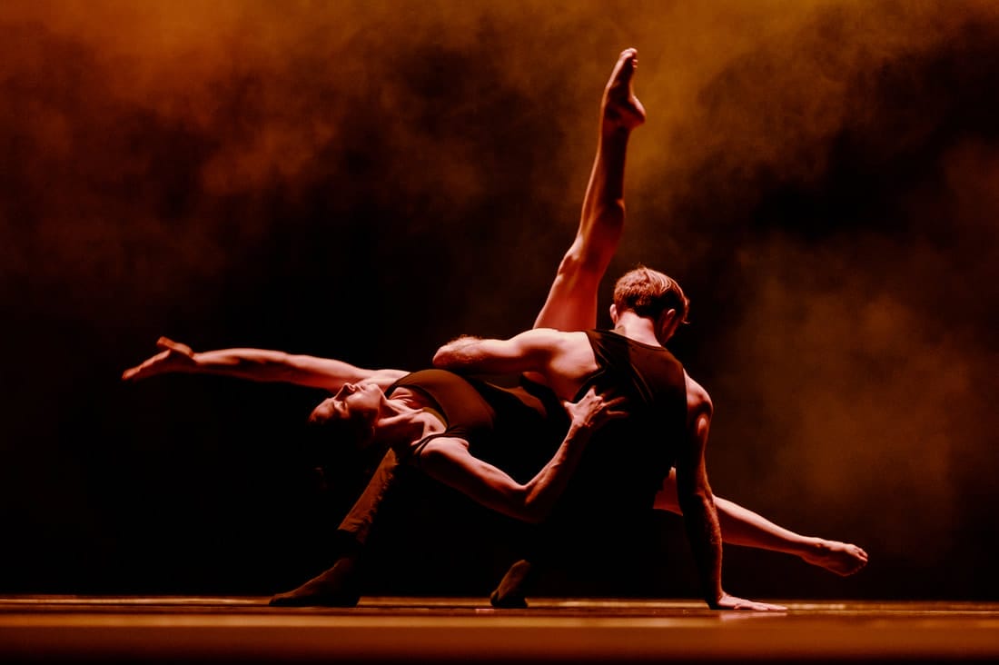 A pair of dancers wearing black vests dance with their bodies intertwined. An orange light and some smoke is around them