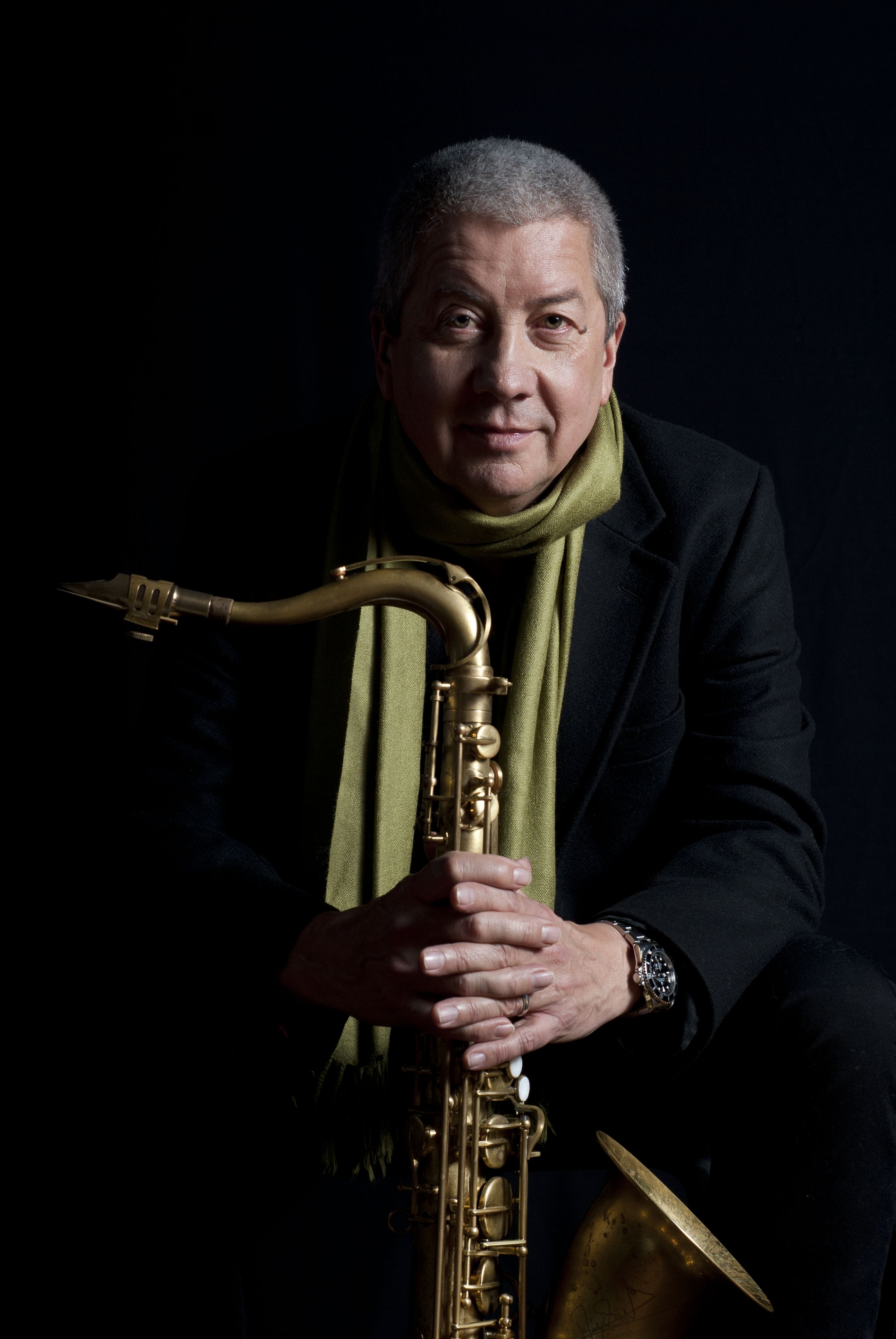 A man stands holding his saxophone - wearing a green scarf.
