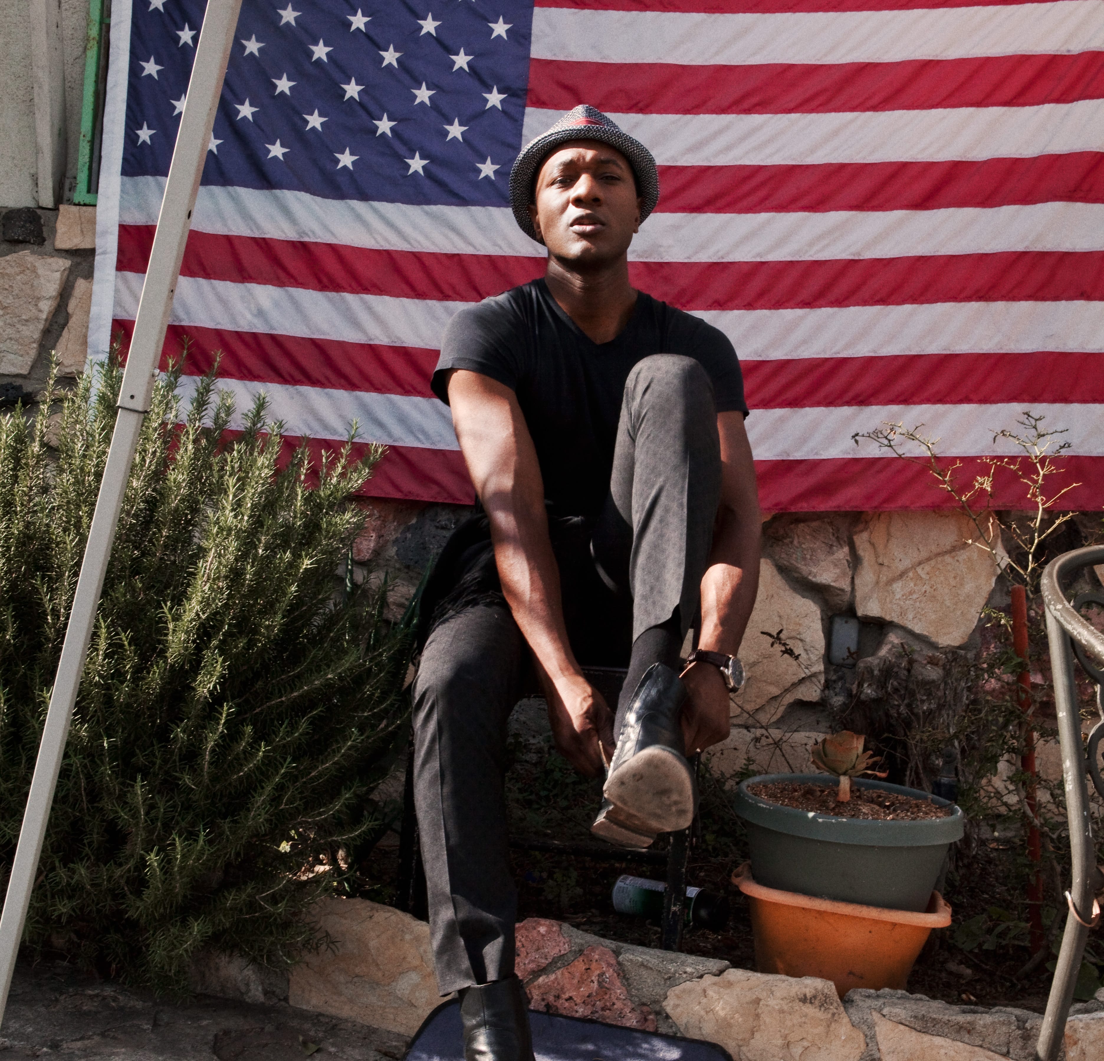 Musician Aloe Blacc skits on a doorstep with an American flag behind him.