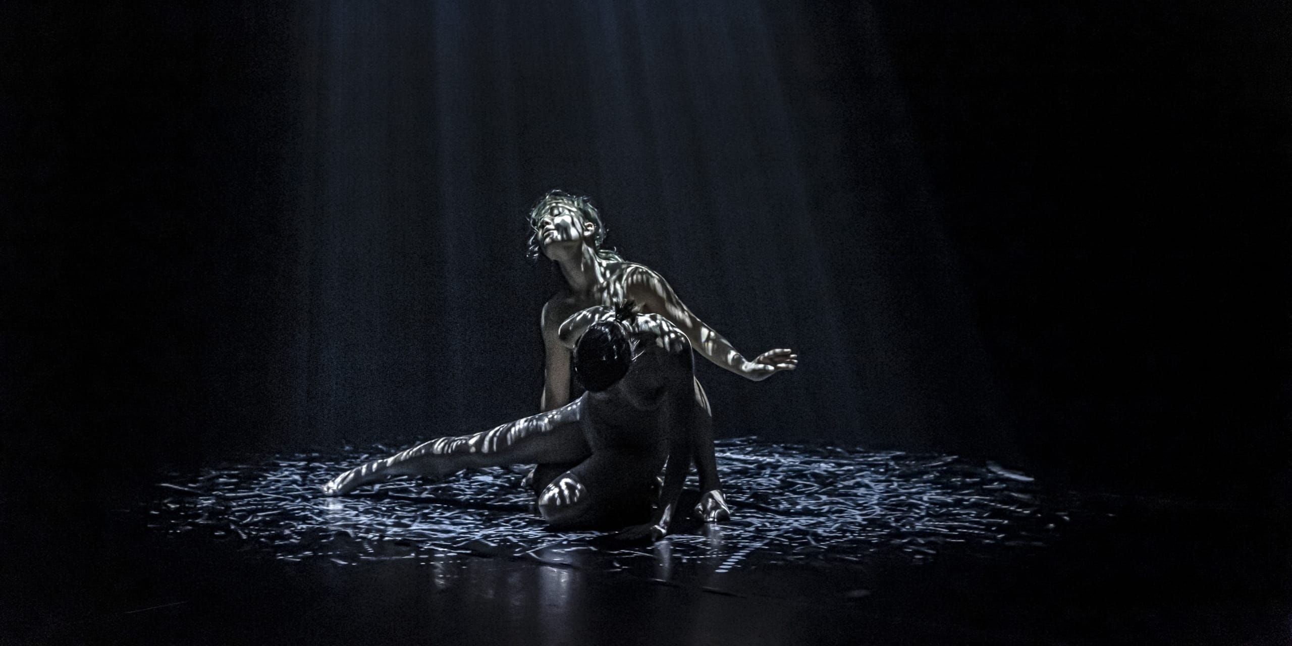 Two female dancers are performing on an all black stage with some patterned light on them giving the effect of rain