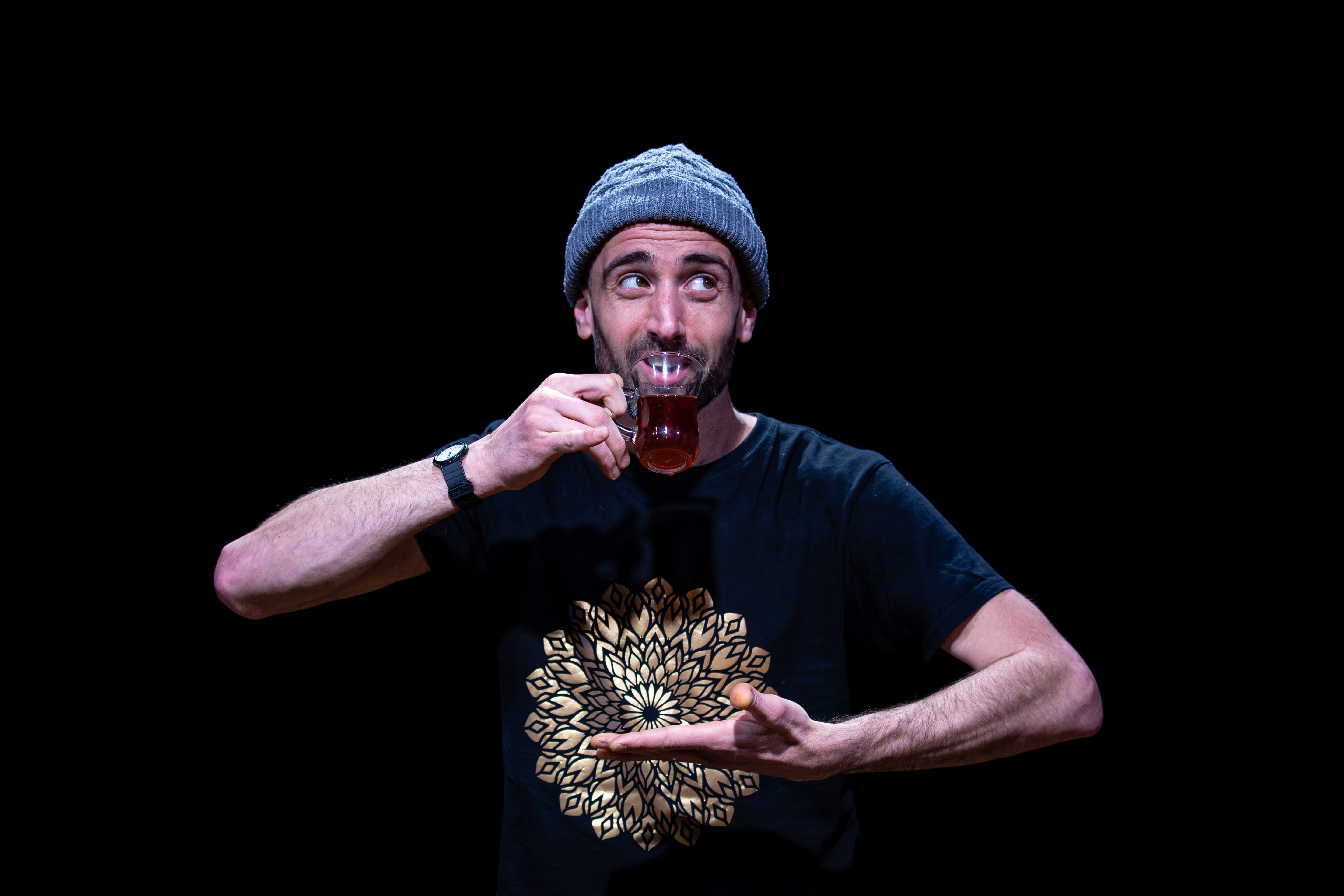 A man on a black bakcground wearing a knitted hat drinks tea from a glass